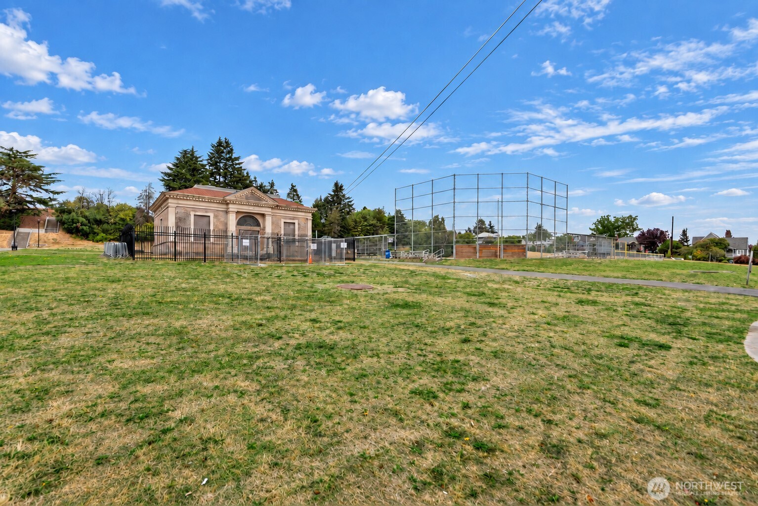 Grassy recreation field with a fenced ball diamond, small building, and homes beyond.