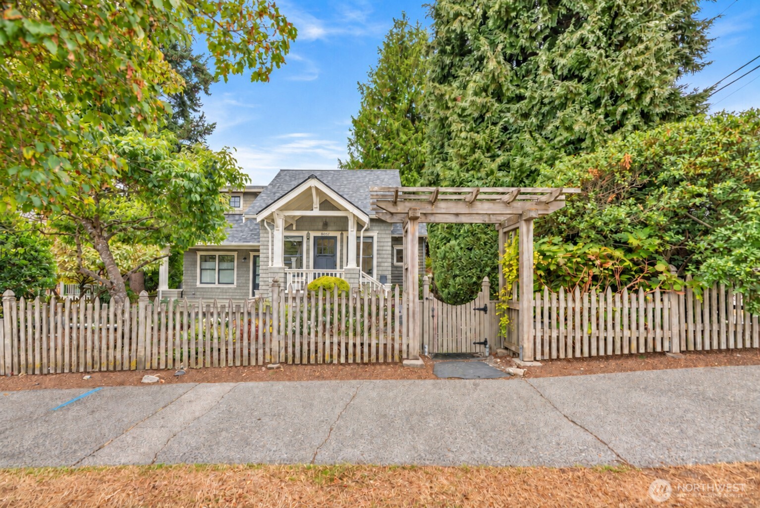 Gray-shingled home with a covered porch behind a white picket fence and wooden pergola gate.