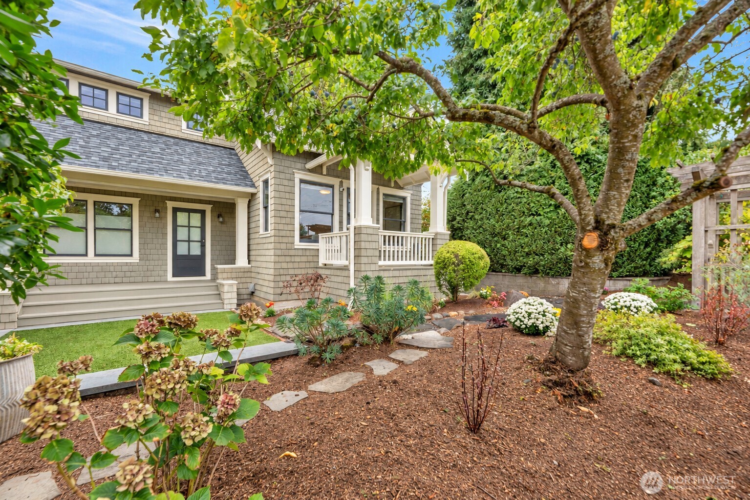 Shingle-sided home with a covered porch, framed by a mature tree and landscaped garden.
