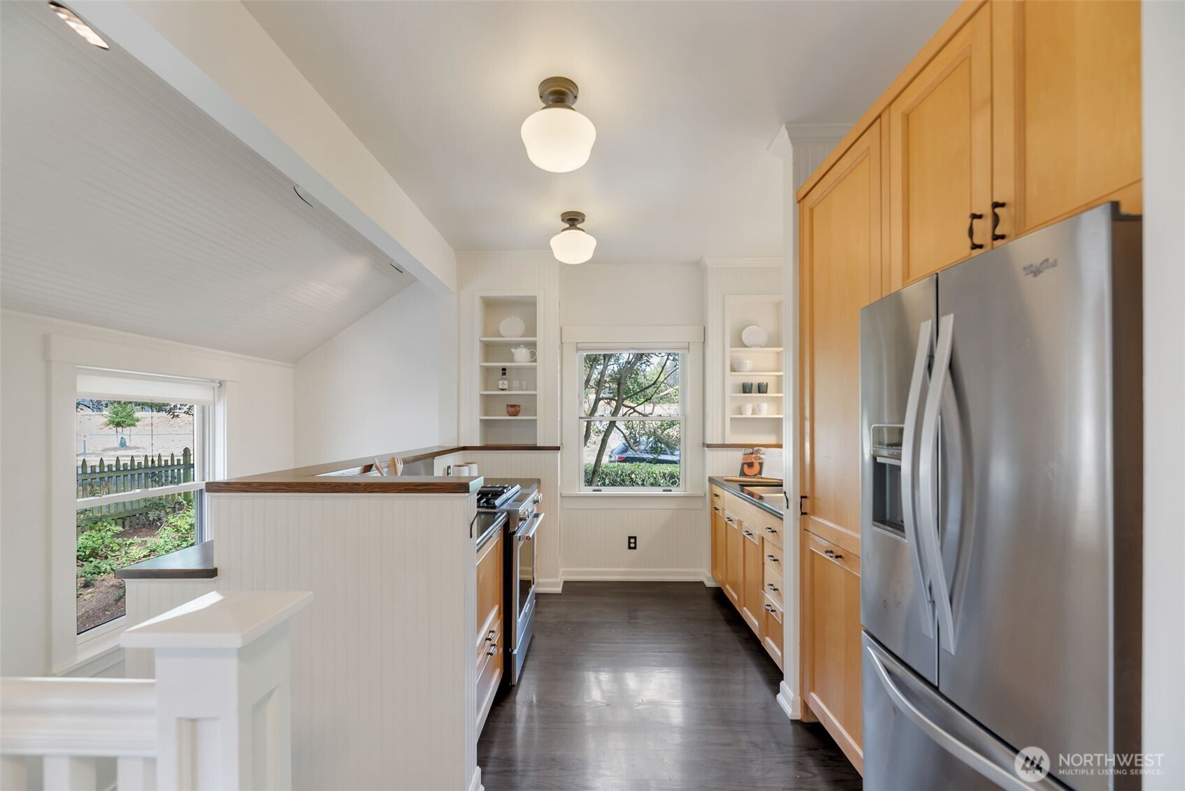 Bright galley kitchen with light wood cabinets, stainless refrigerator, dark flooring, and a raised breakfast counter.