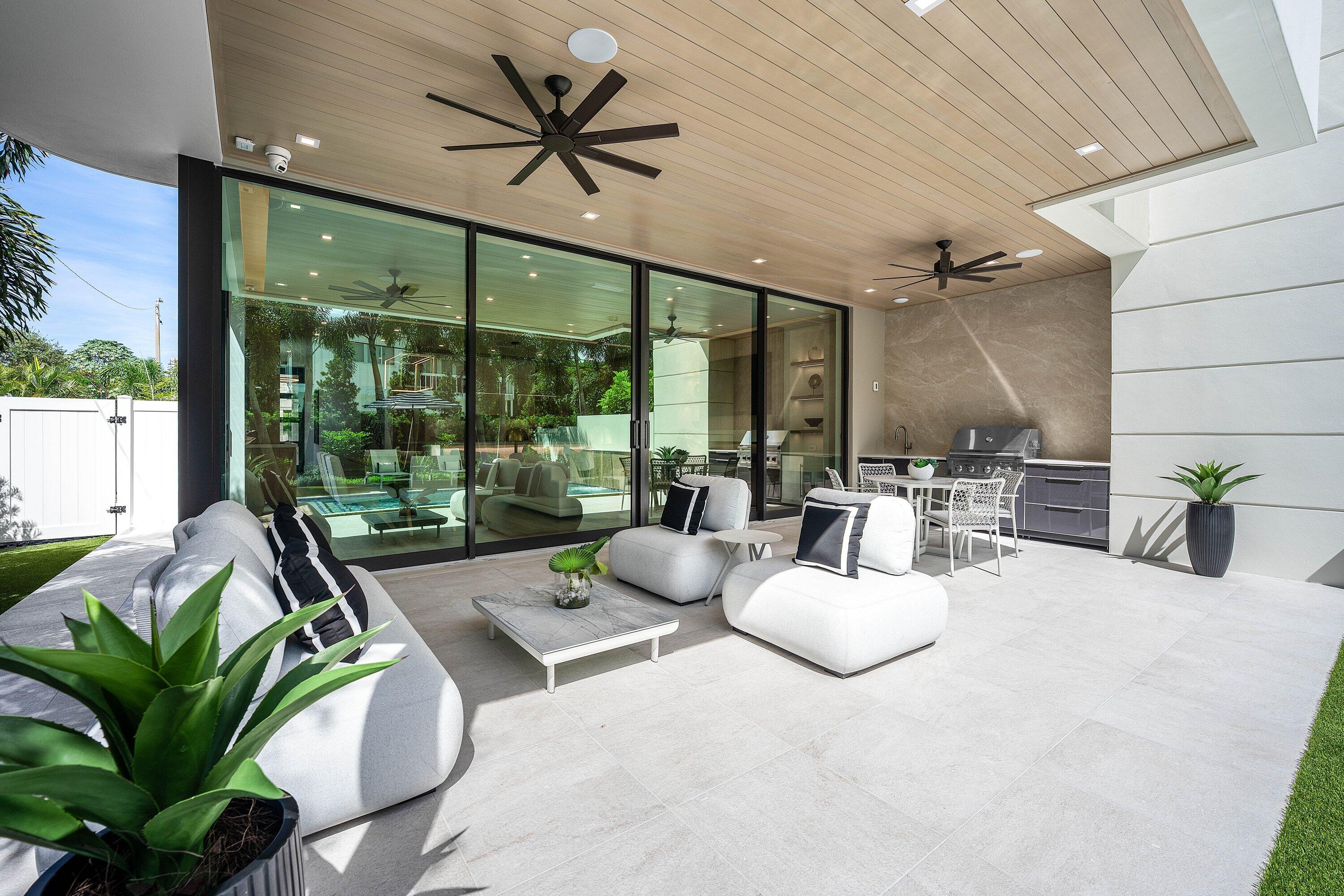 Covered patio with white lounge seating, ceiling fans, and an outdoor grill beside glass doors overlooking a pool.