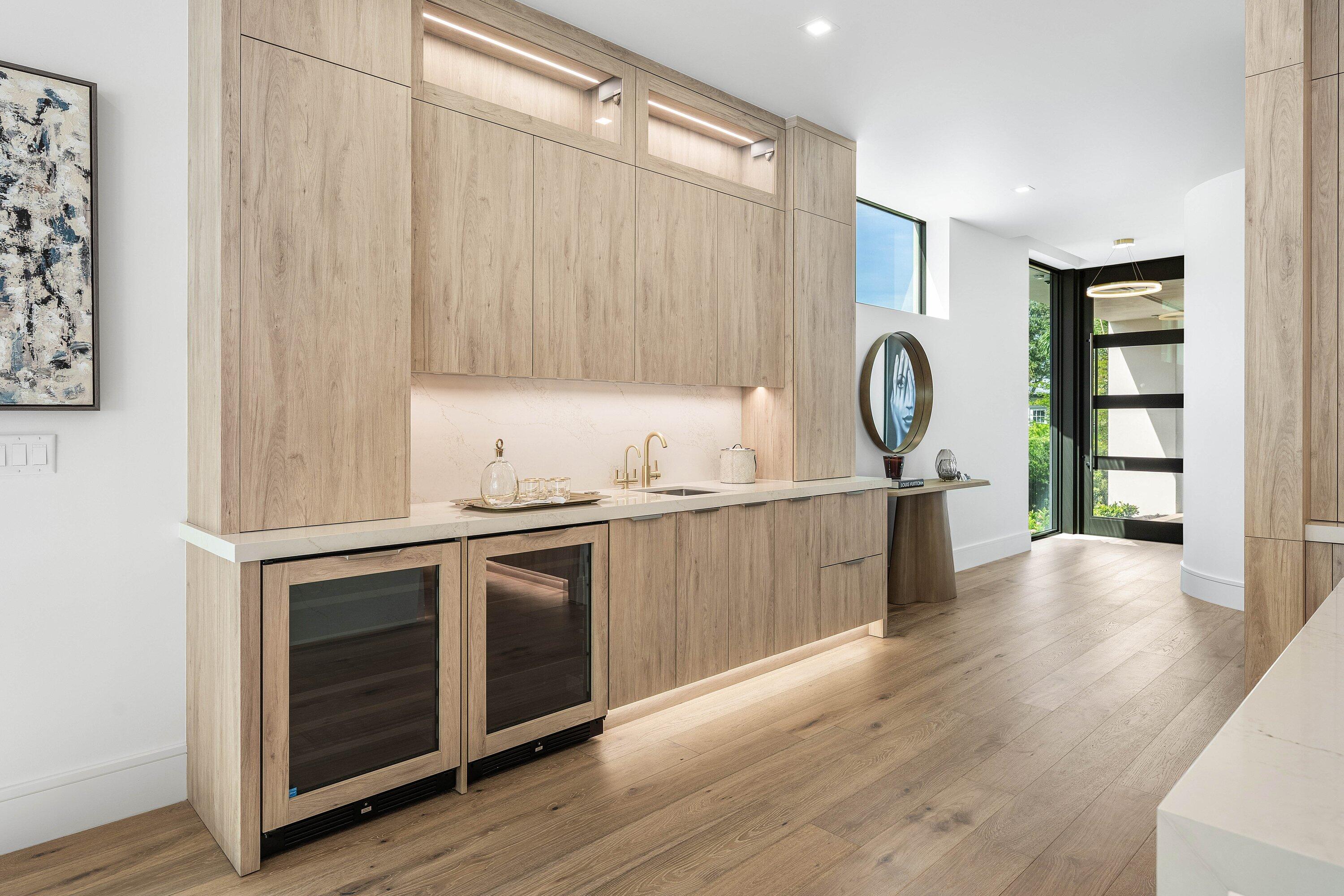 Modern oak kitchen bar with brass faucet, stone backsplash, and two glass-front wine refrigerators.