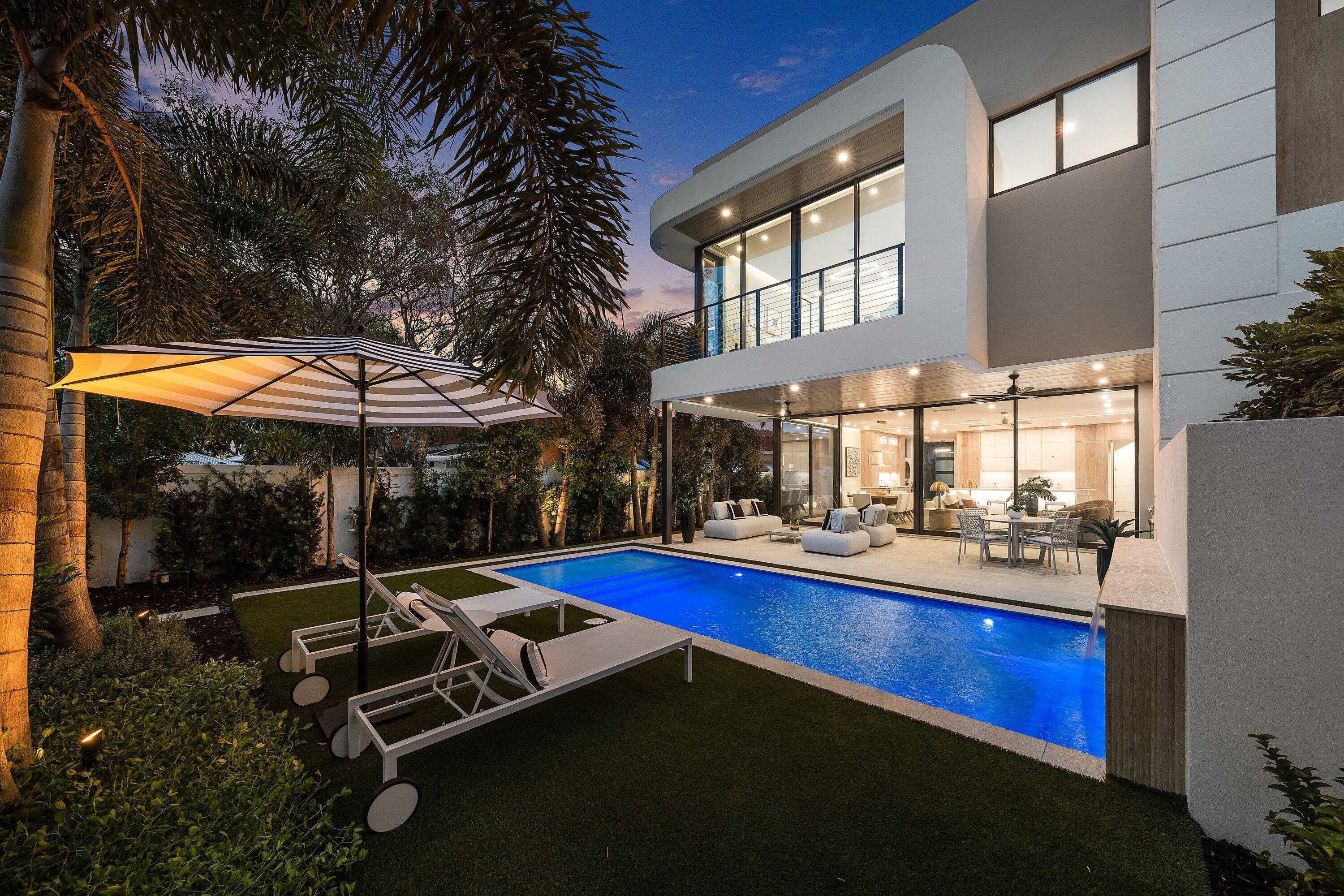 Modern home and illuminated backyard pool at dusk, with glass walls, tropical landscaping, and outdoor seating.