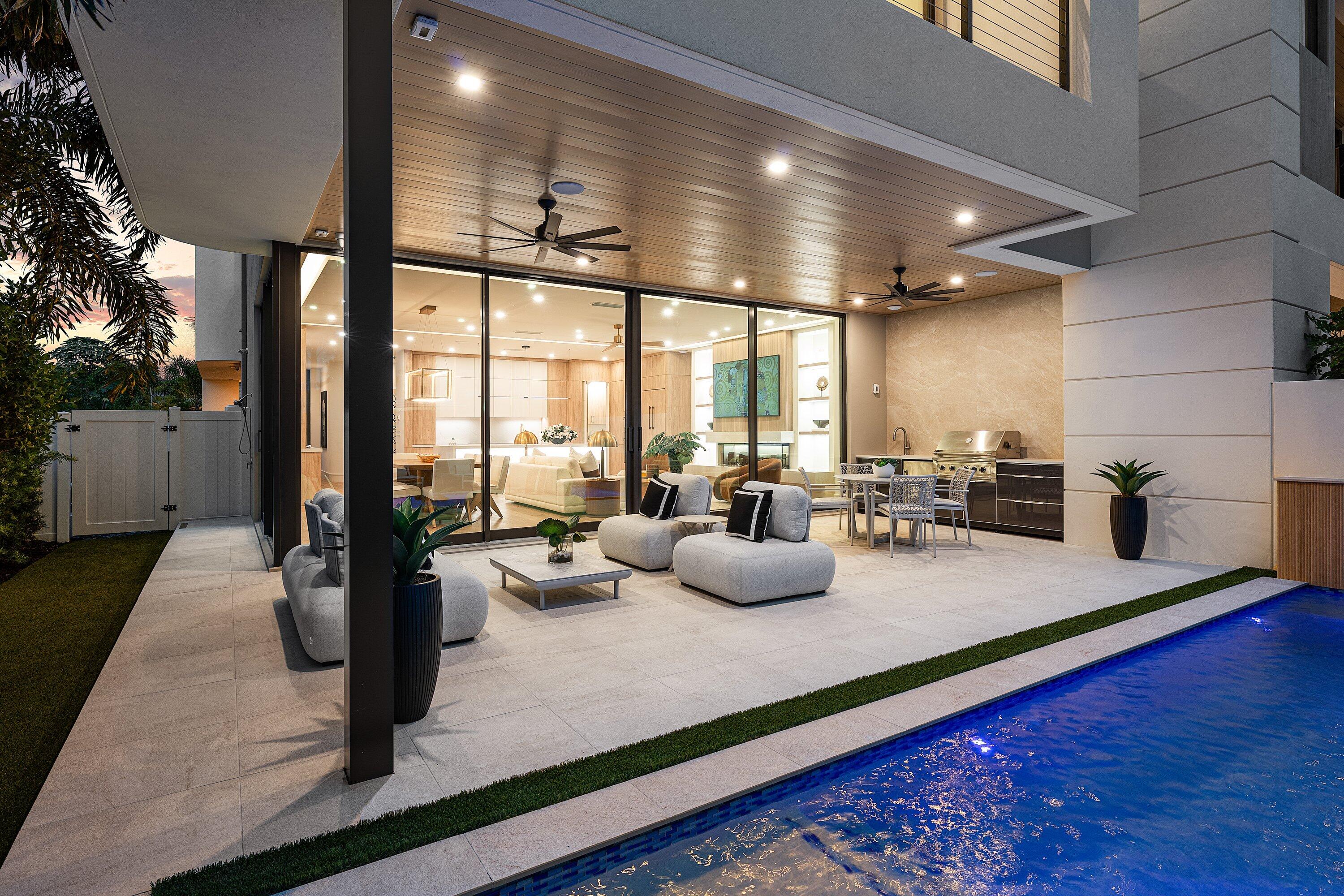 Covered patio with modern lounge seating and outdoor kitchen beside an illuminated swimming pool at dusk.