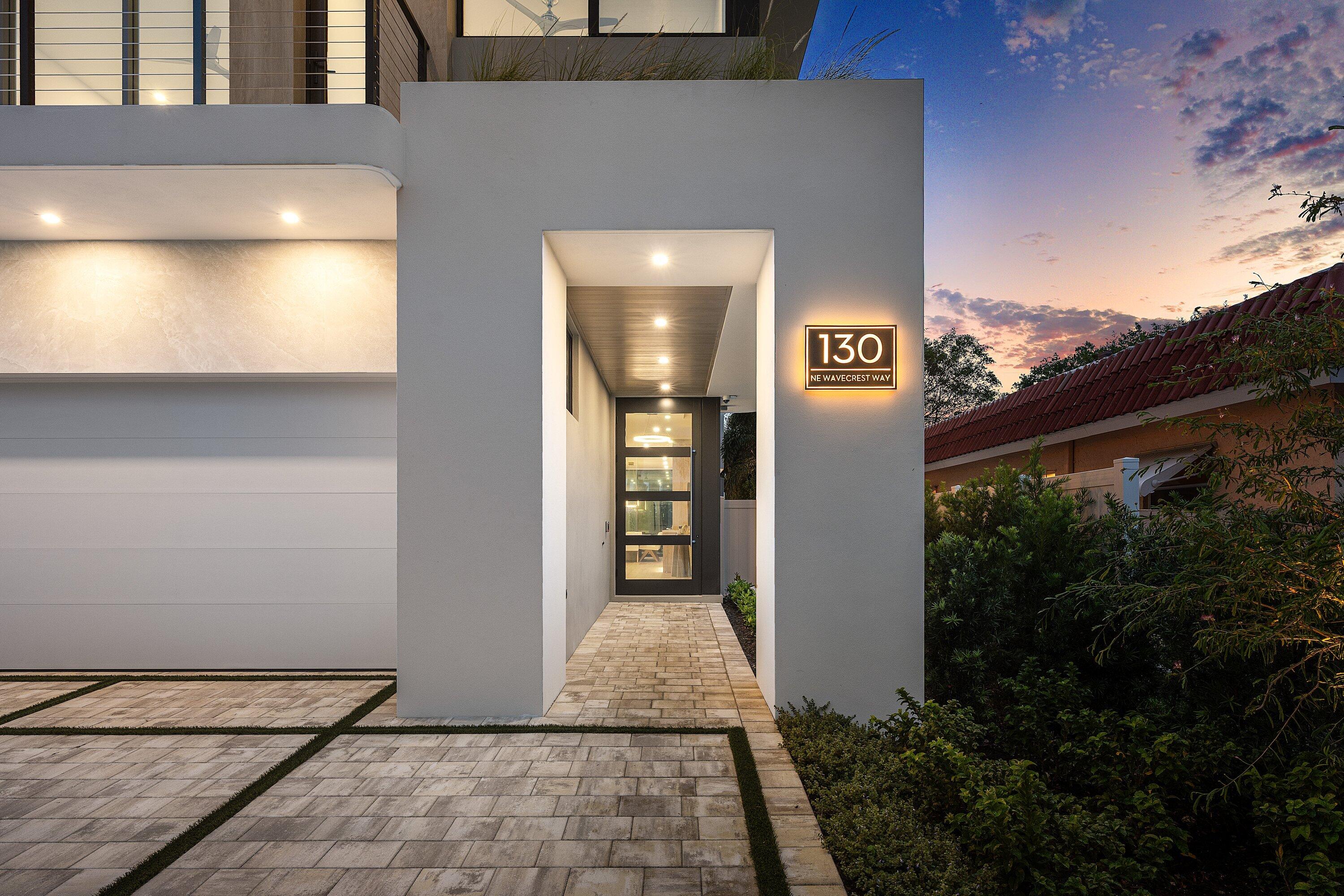 Modern home entrance with a recessed glass door and illuminated 130 NE Wavecrest Way sign at dusk.