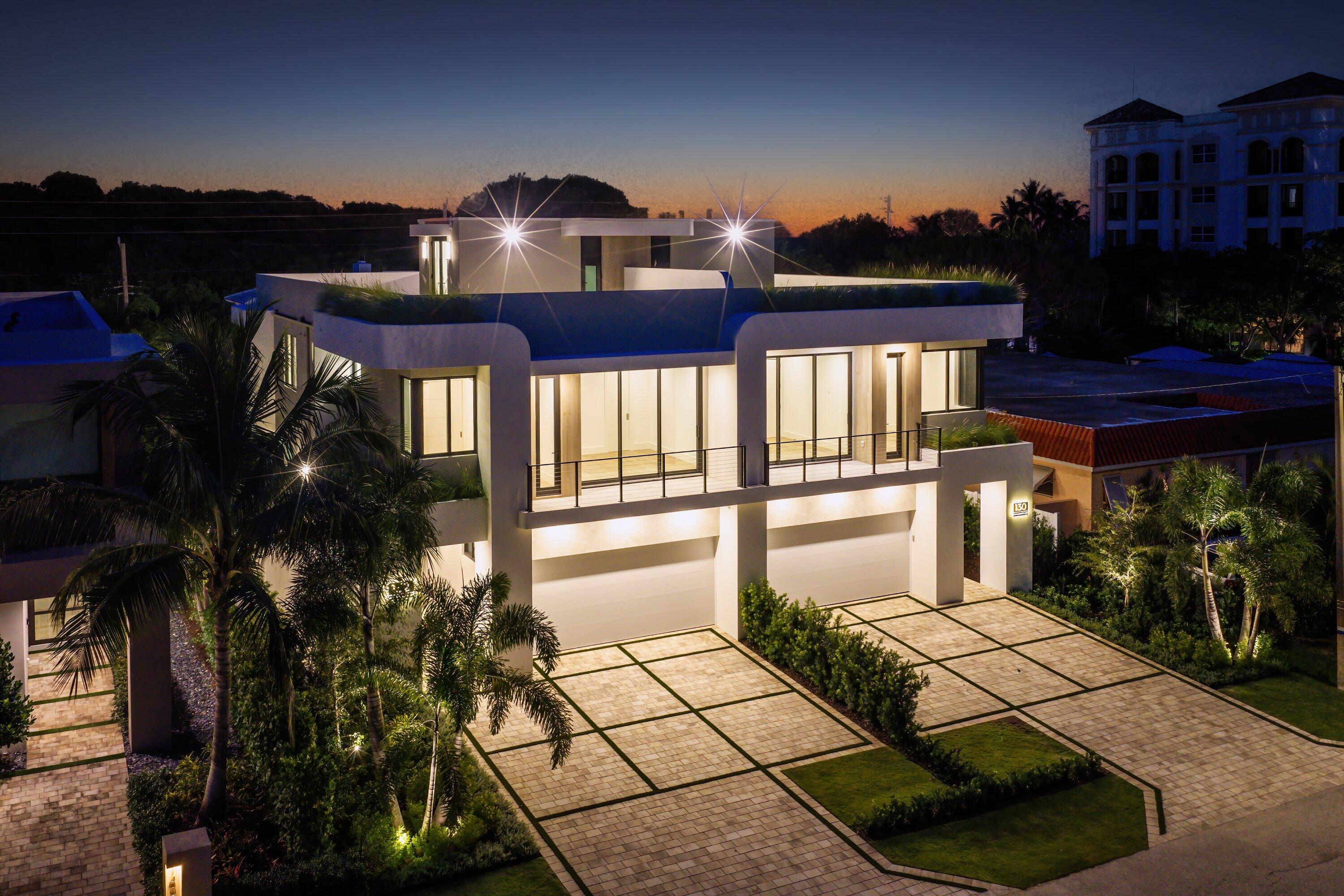 Modern two-story home at dusk with an illuminated balcony, palm landscaping, and a geometric-paver driveway.