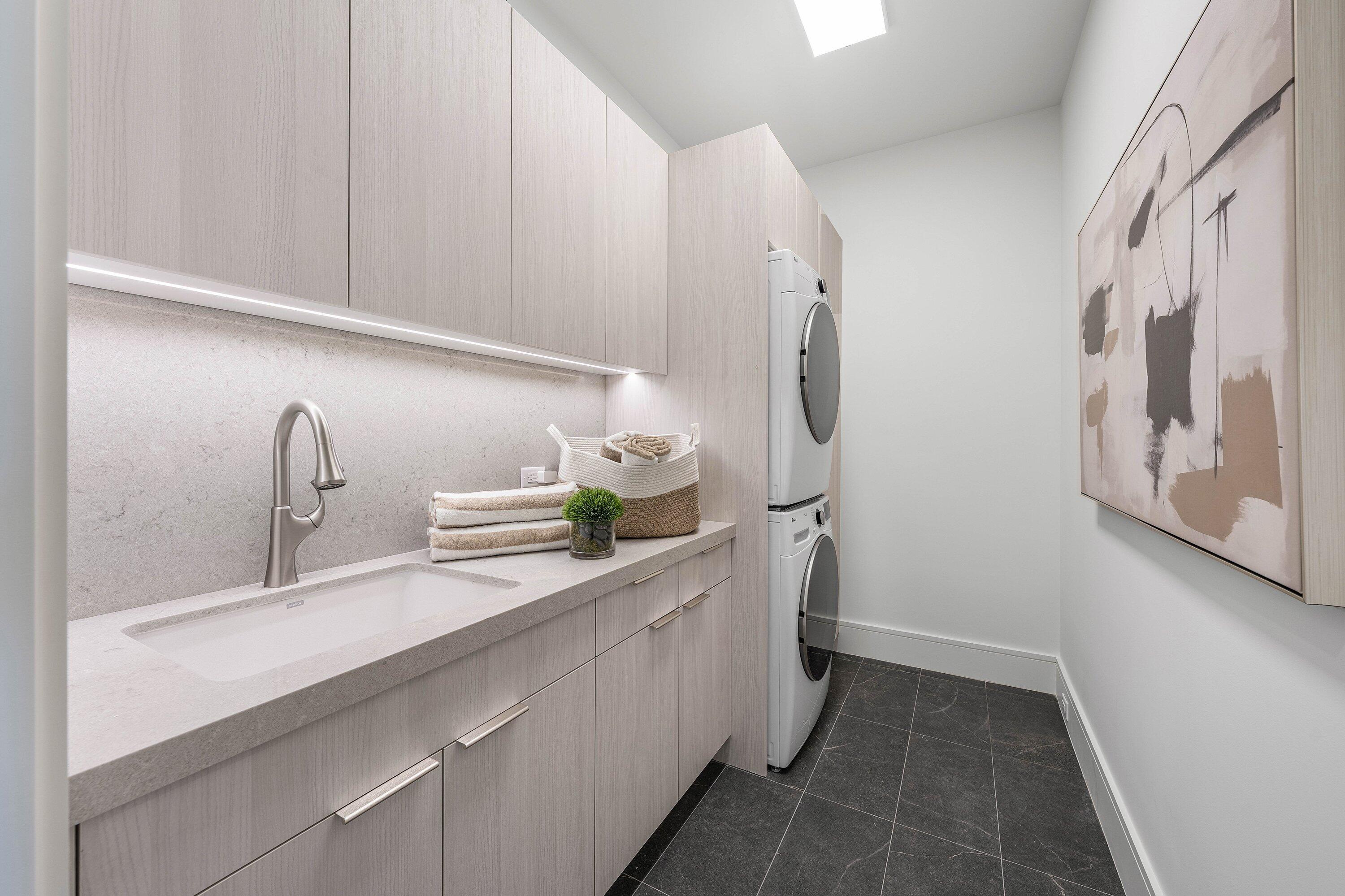 Modern laundry room with light wood cabinets, an integrated sink, and stacked washer and dryer.