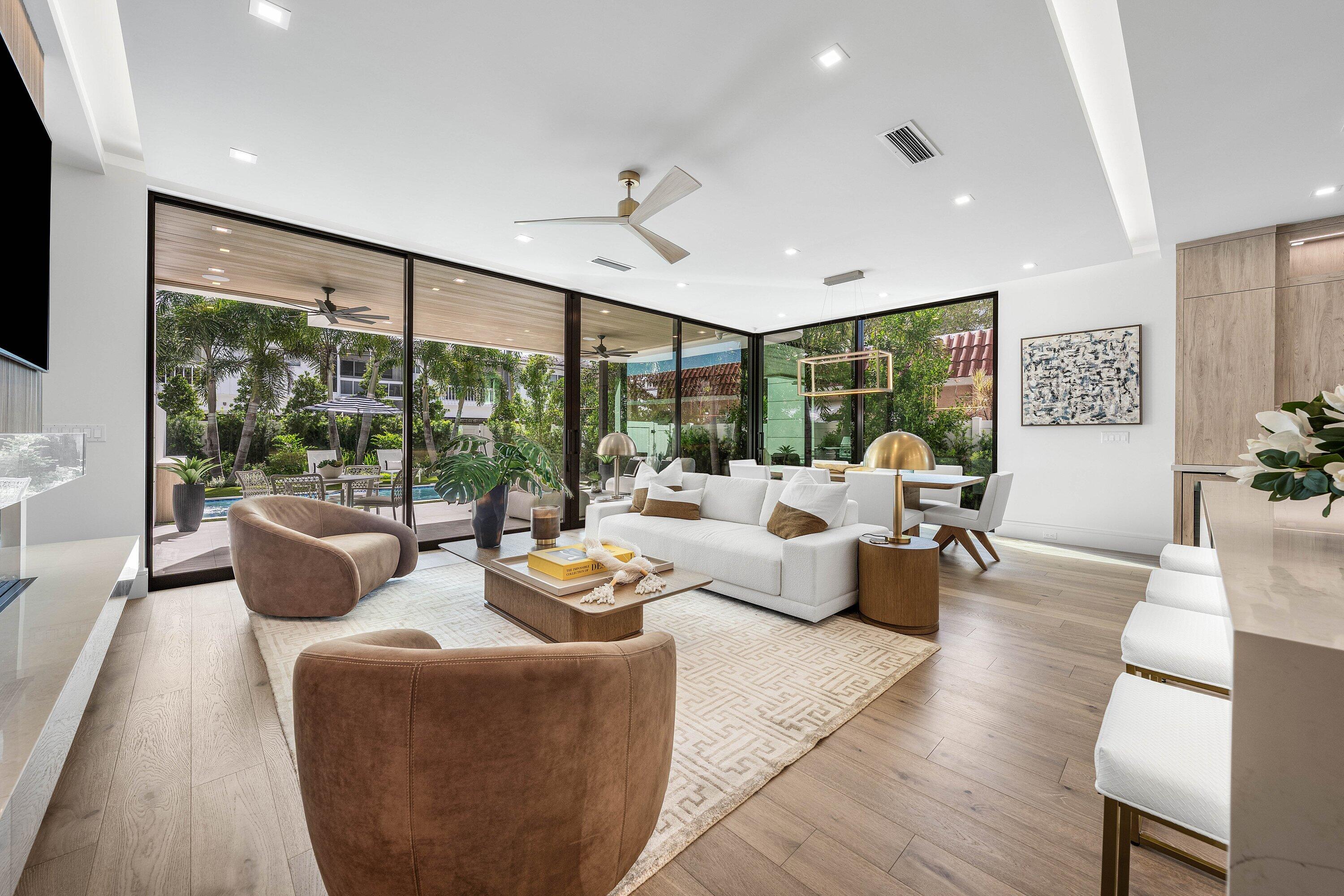 Modern living room with white sofas and floor-to-ceiling glass doors overlooking a landscaped patio and pool.