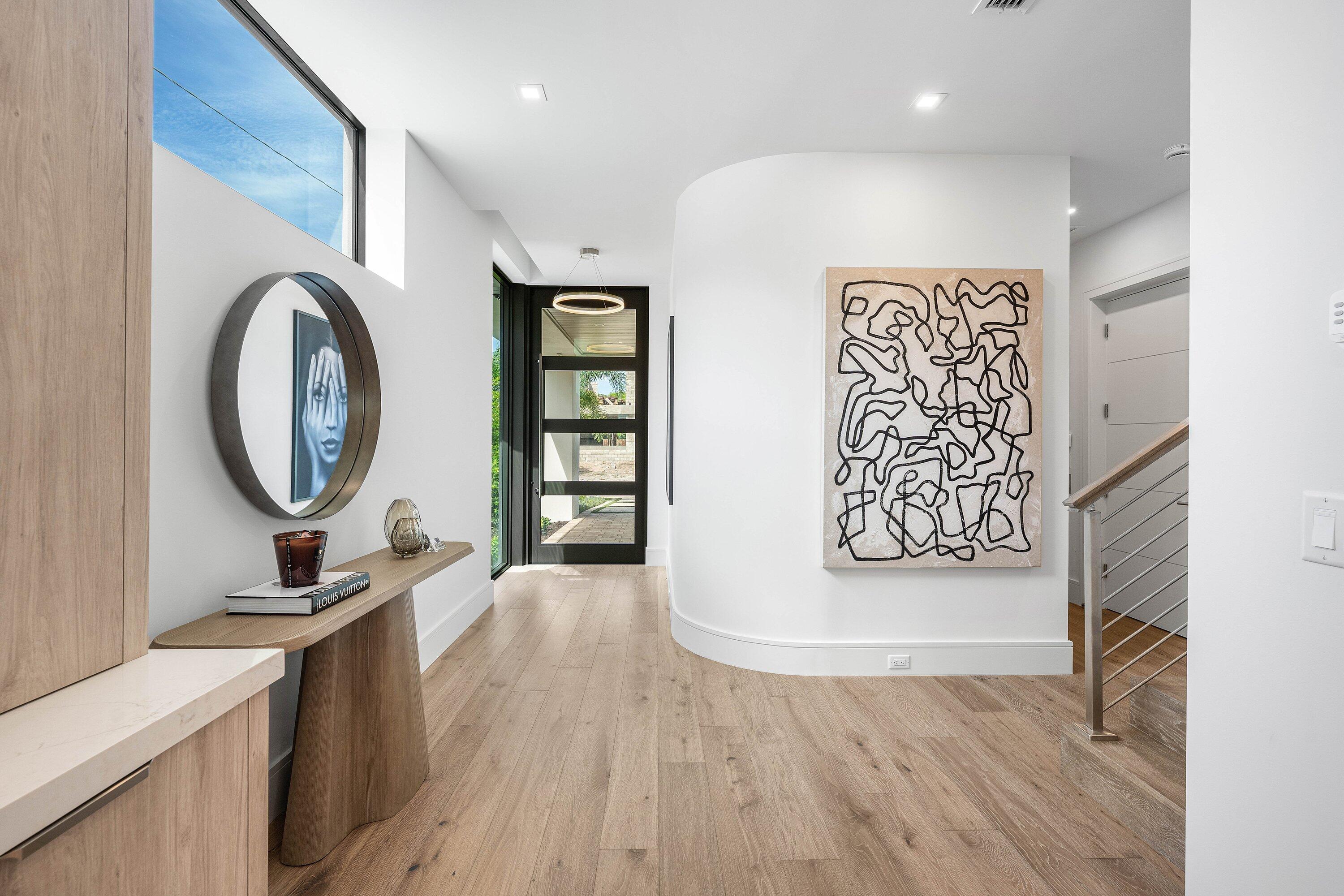 Modern foyer with light wood floors, a curved art wall, glass entry door, and metal-railed staircase.