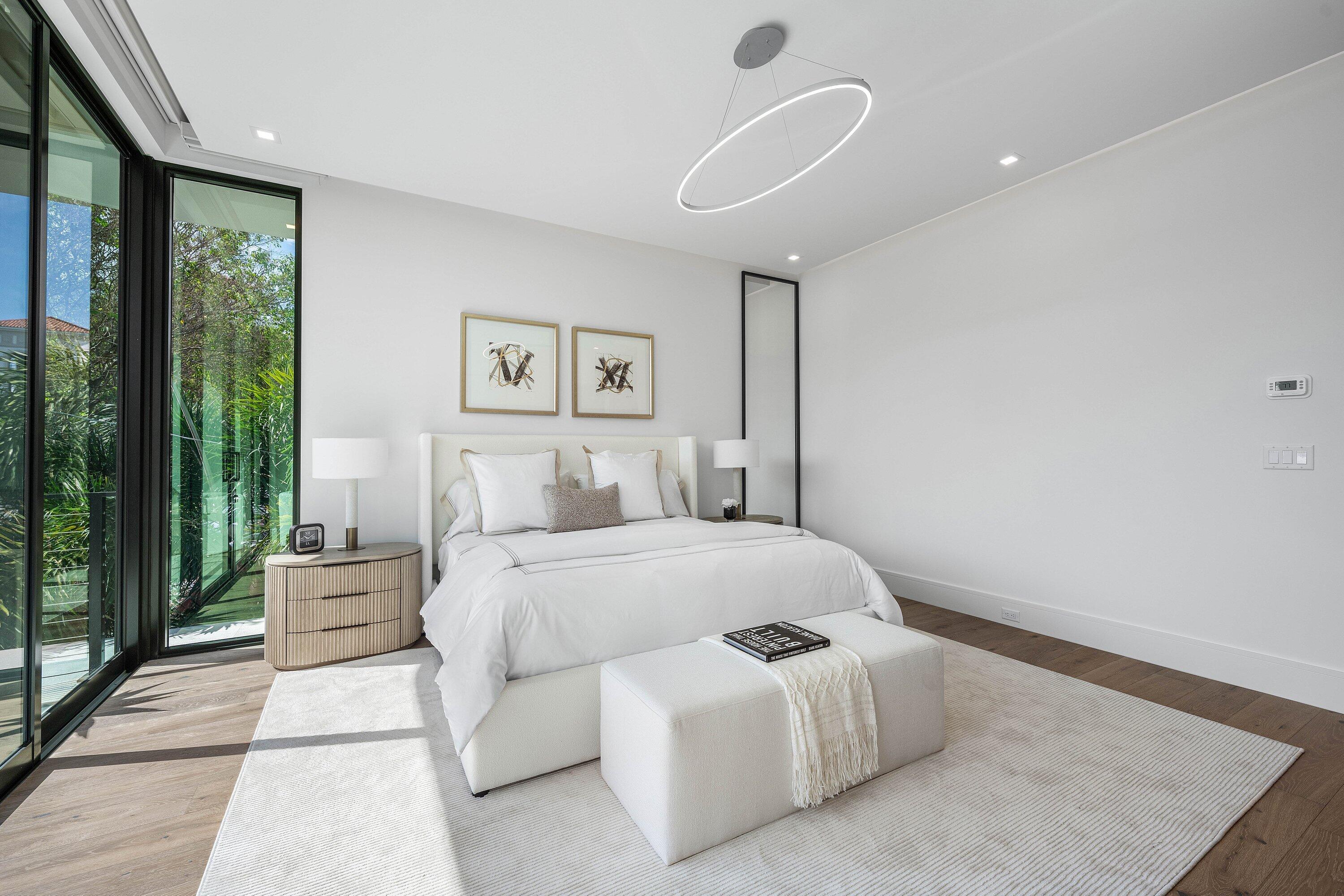 Modern bedroom with a white upholstered bed beside floor-to-ceiling glass doors overlooking greenery.