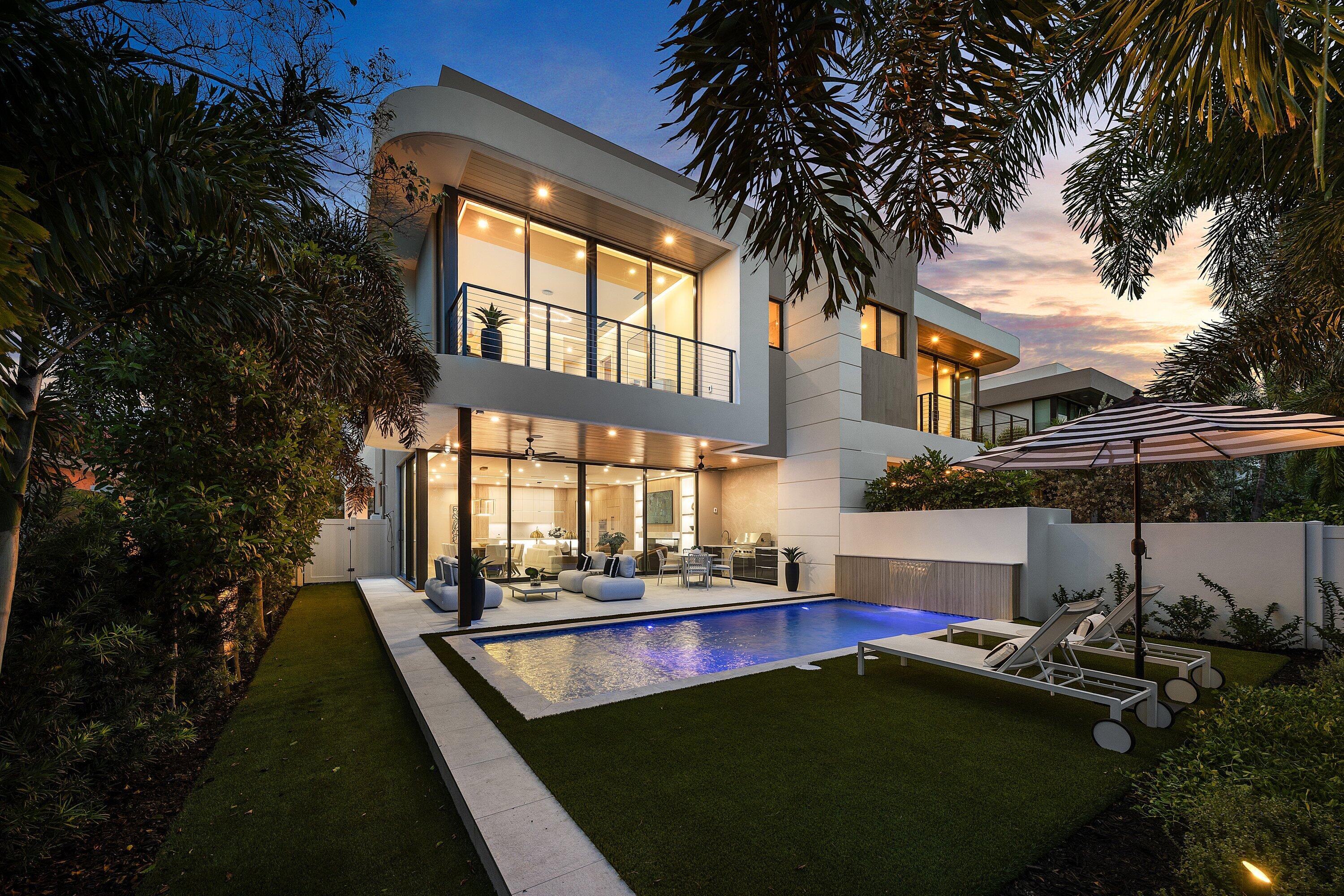 Modern two-story home beside an illuminated pool, with glass walls and a furnished patio at dusk.