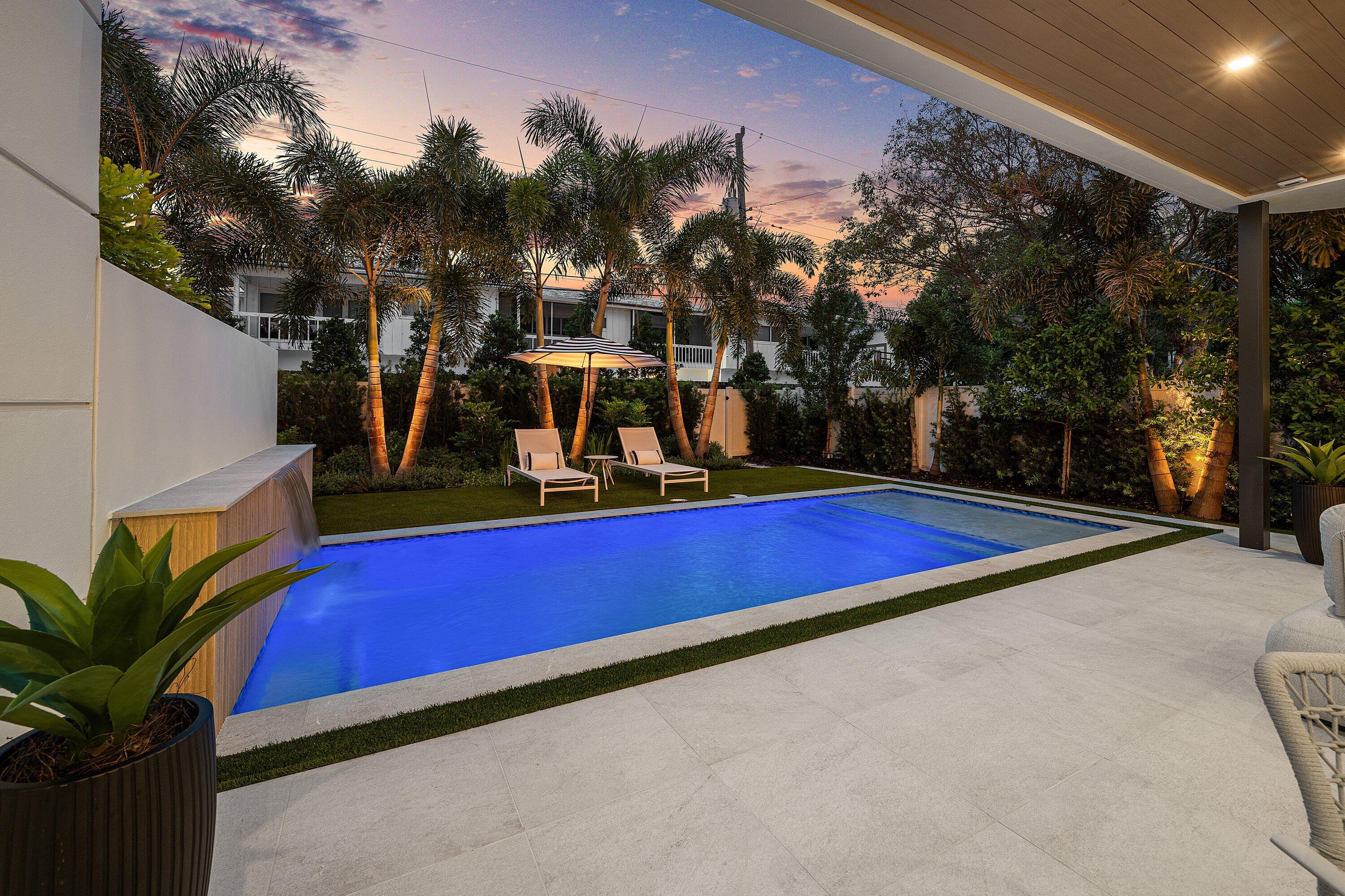 Backyard pool beside a covered patio, with palm trees and lounge chairs beneath a striped umbrella at dusk.