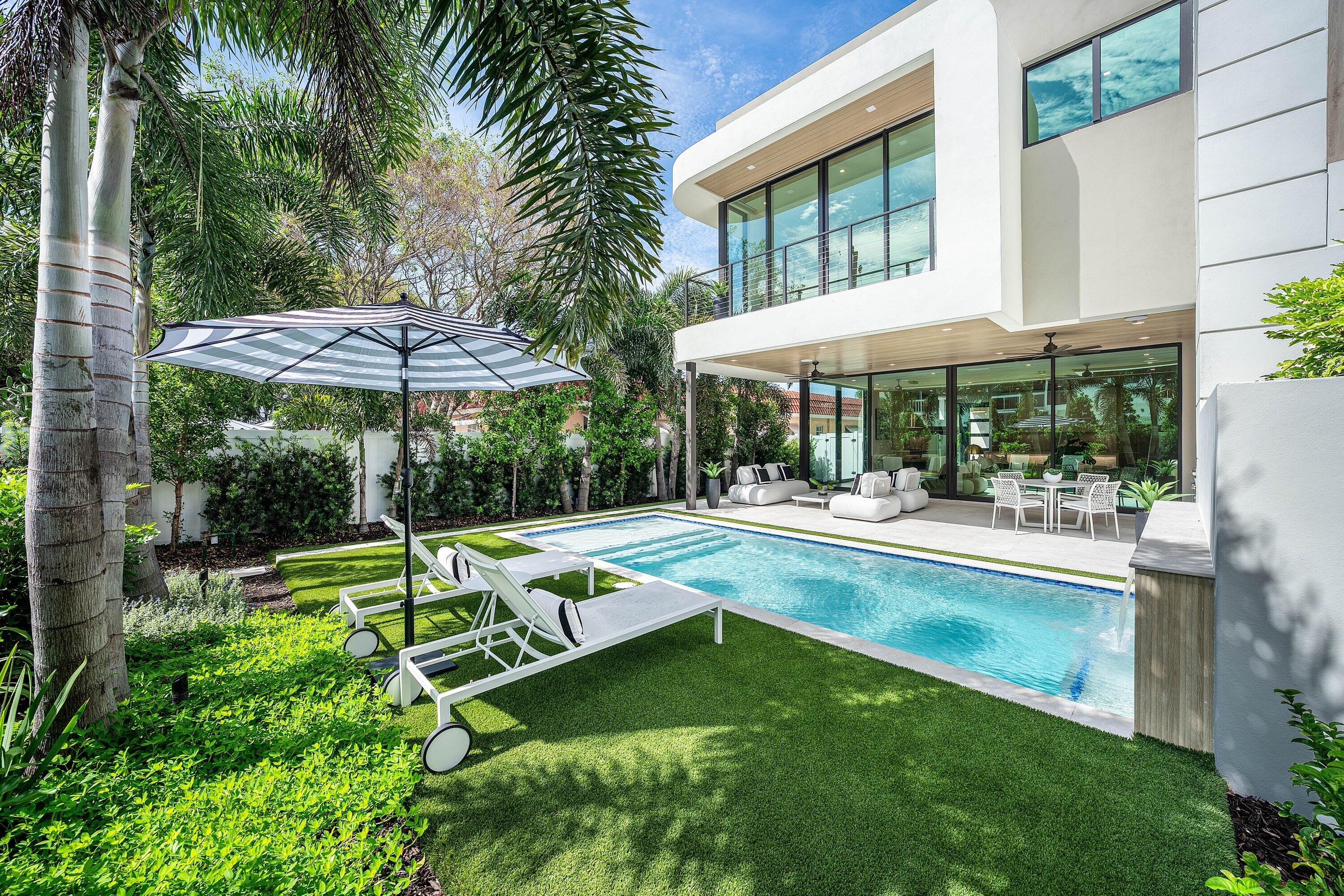 Modern white home with a long pool, furnished patio, and tropical landscaping.