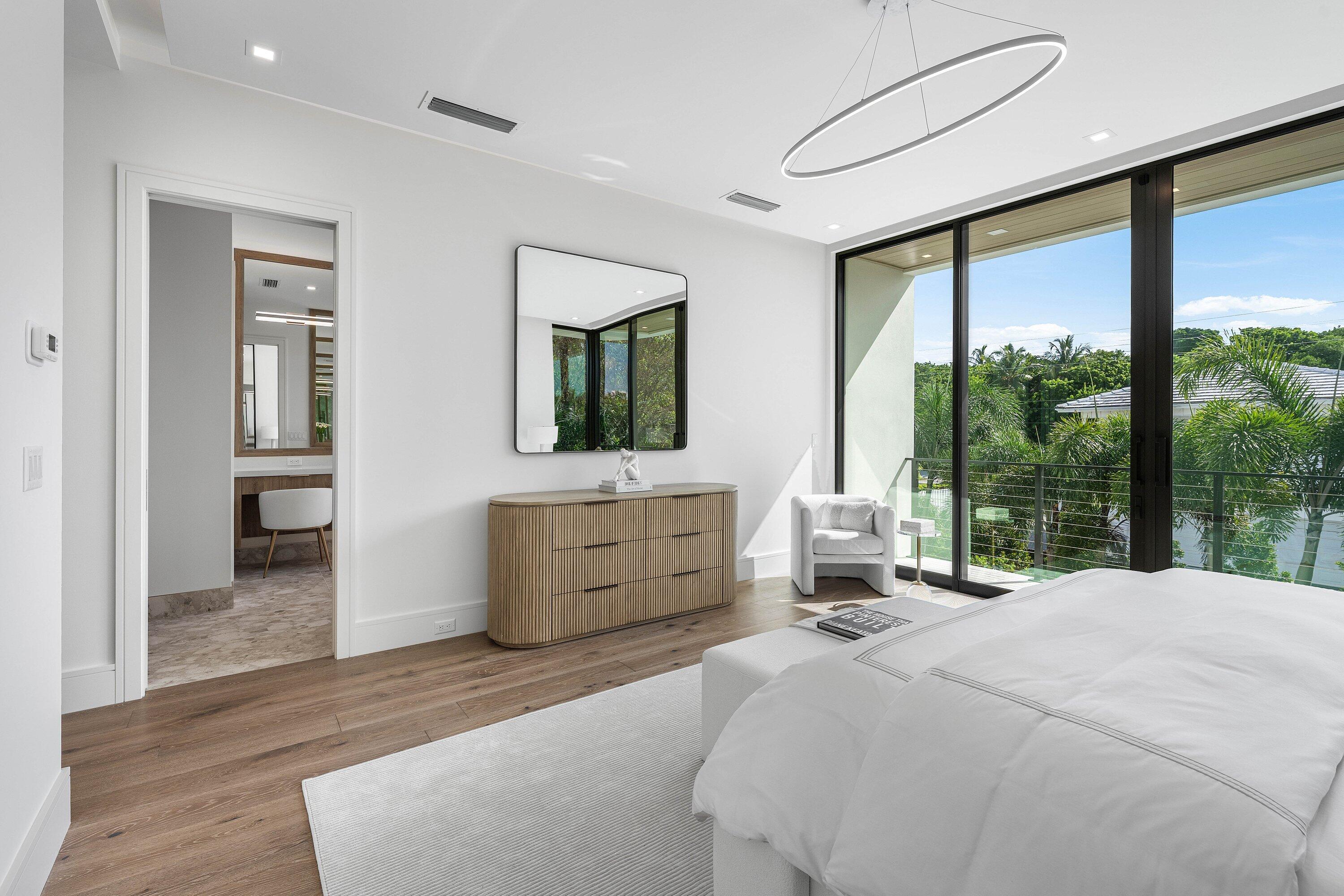 Modern bedroom with a white bed, wood dresser, and sliding doors overlooking palm trees.
