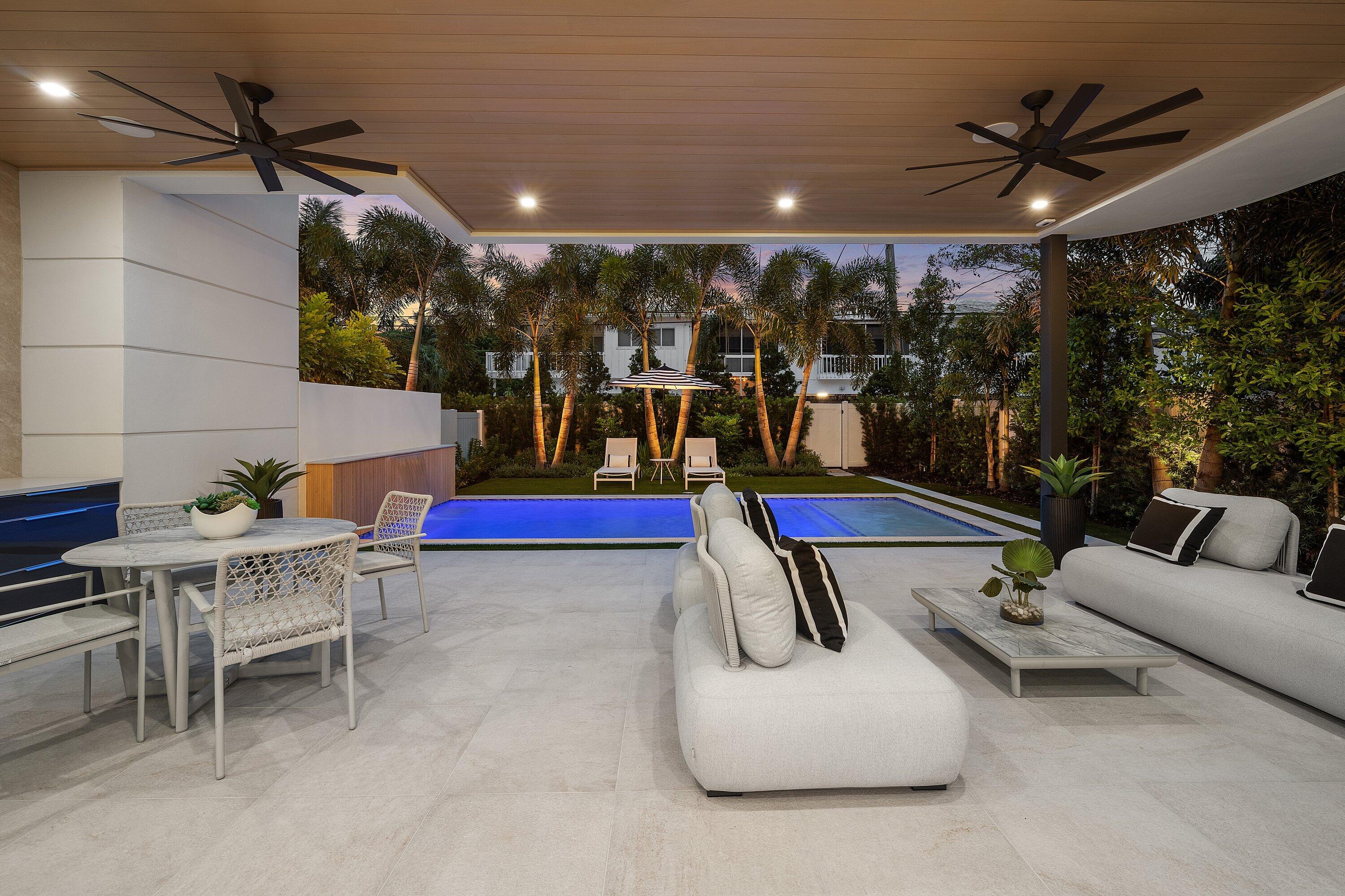 Covered patio with lounge seating and dining area overlooking a blue-lit pool and palm trees.