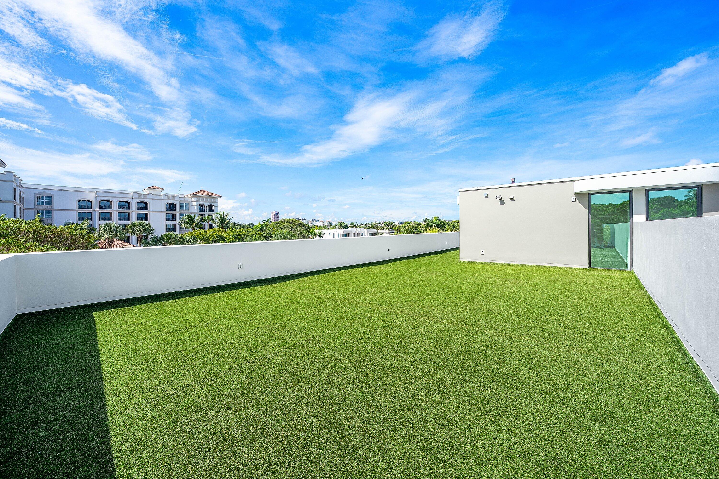 Rooftop terrace with green turf, white walls, and views of palm trees and neighboring buildings.
