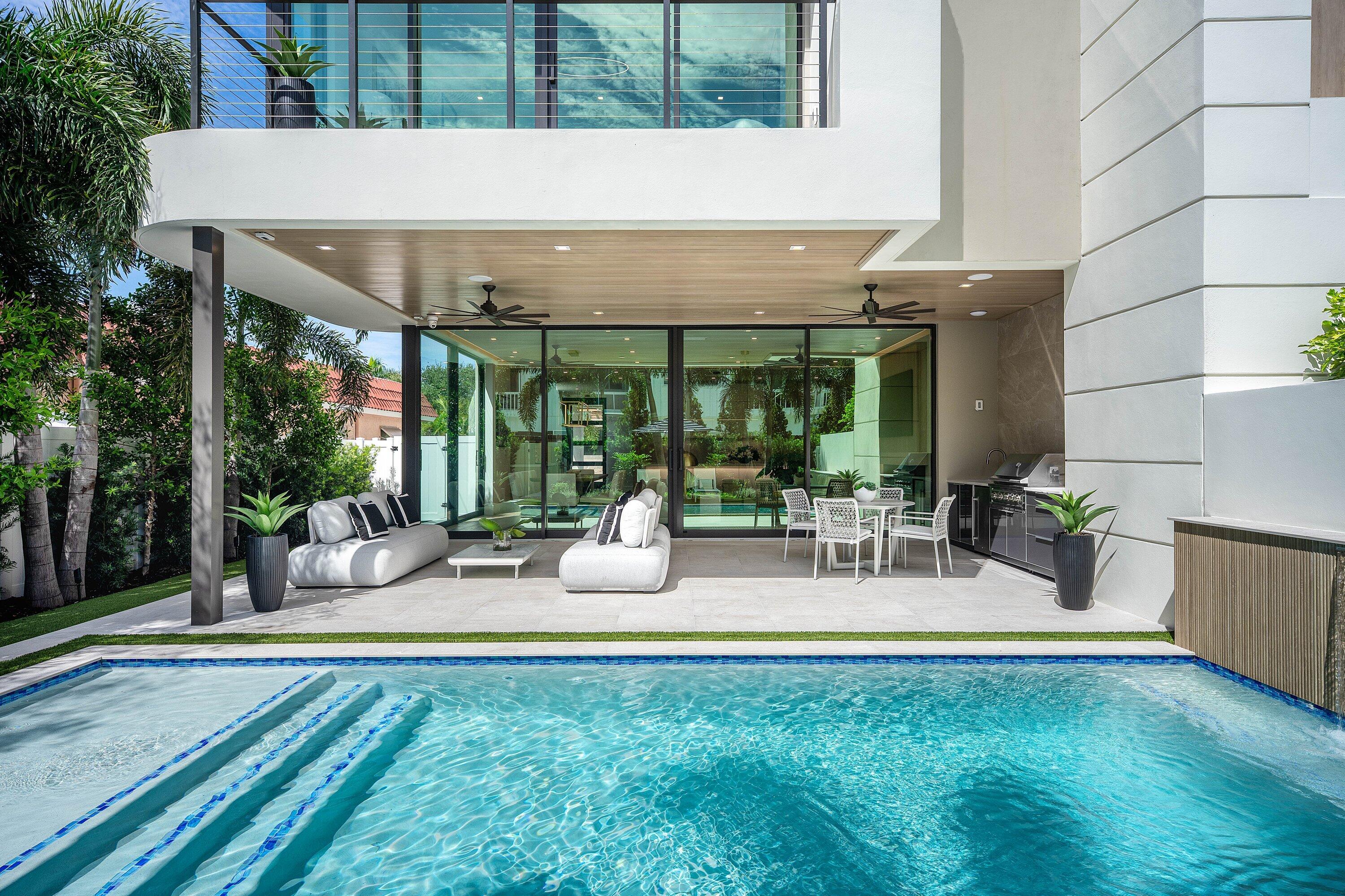 Swimming pool beside a covered patio with white lounge seating, outdoor dining, and a built-in kitchen.