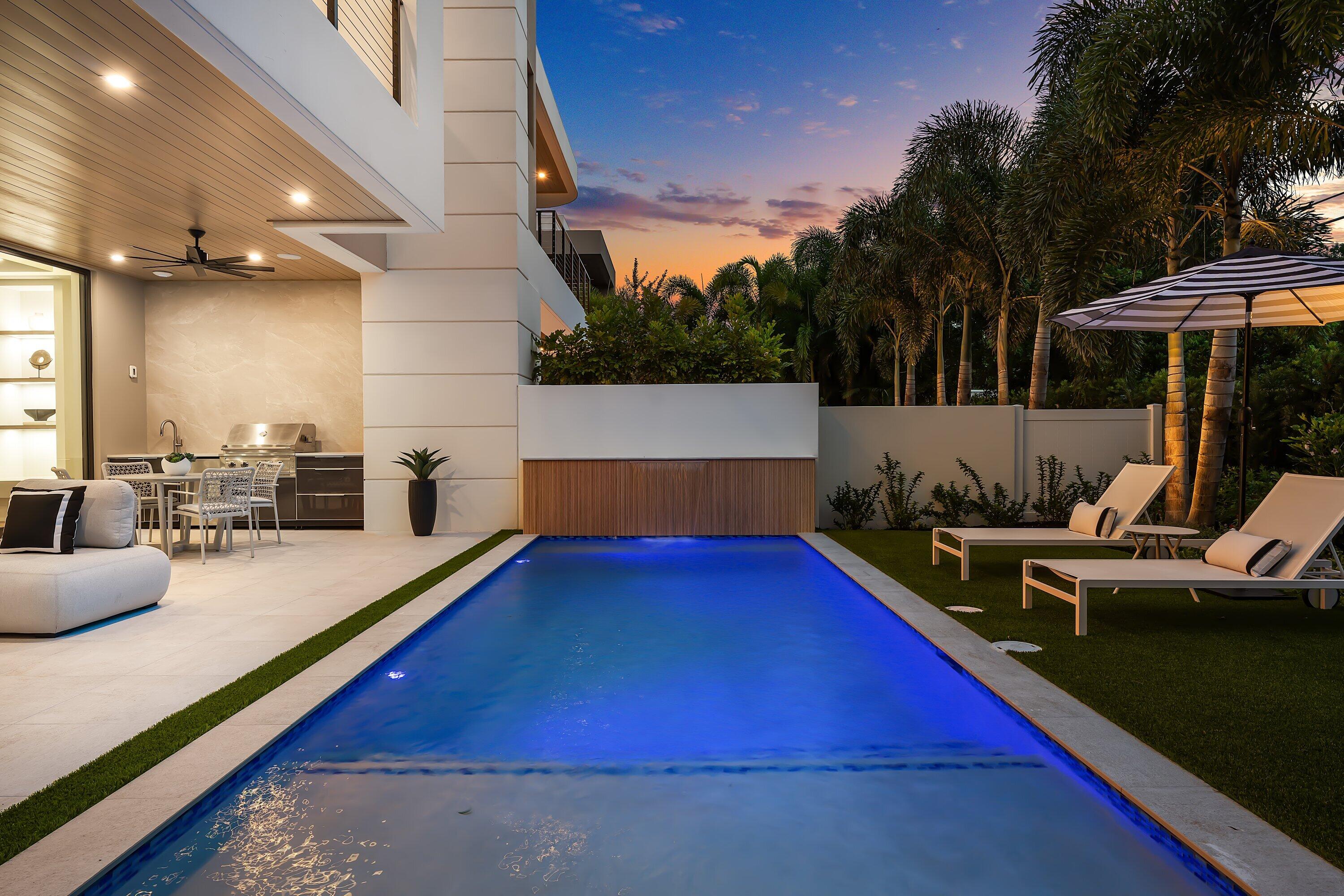 Modern home patio with a long pool, outdoor kitchen, and palm-lined lounge area at dusk.