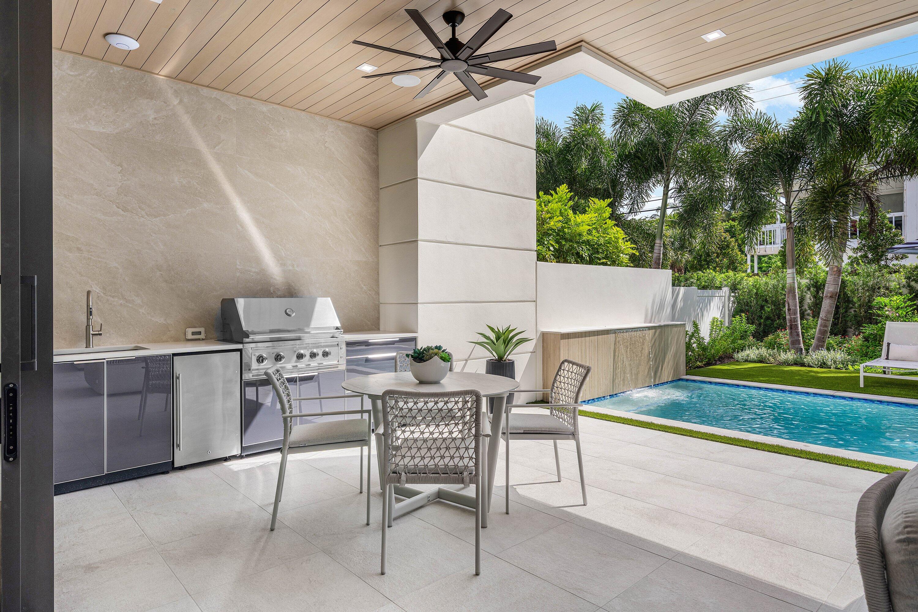 Covered patio with an outdoor kitchen and dining set beside a pool and palm trees.