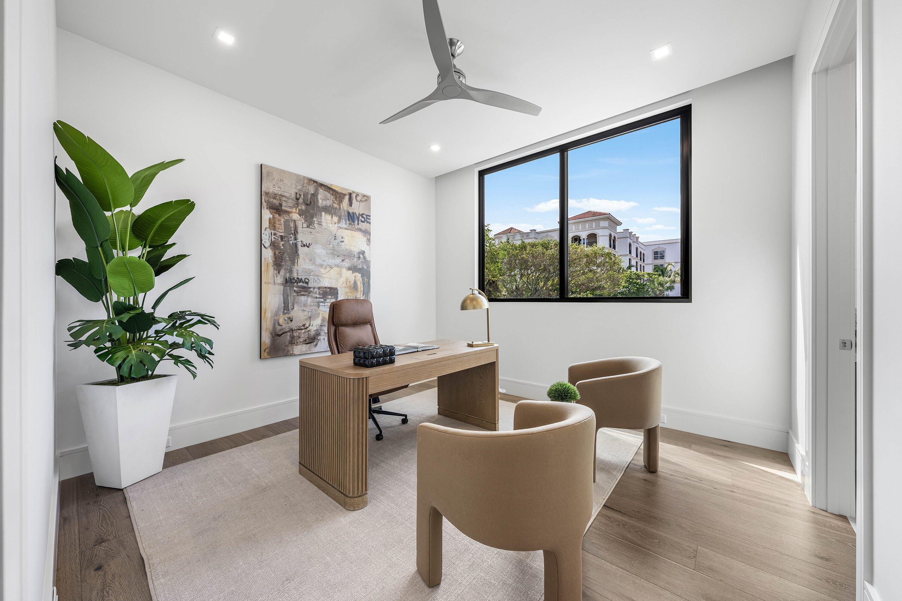Furnished home office with a wood desk, tan chairs, and a large window overlooking trees.