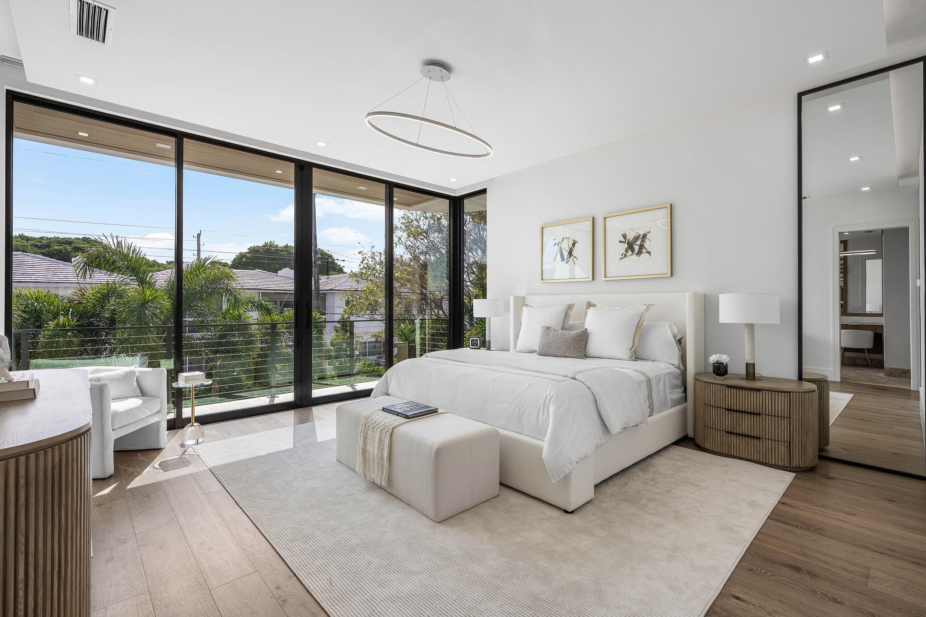 Staged bedroom with a white bed and floor-to-ceiling glass doors overlooking trees.