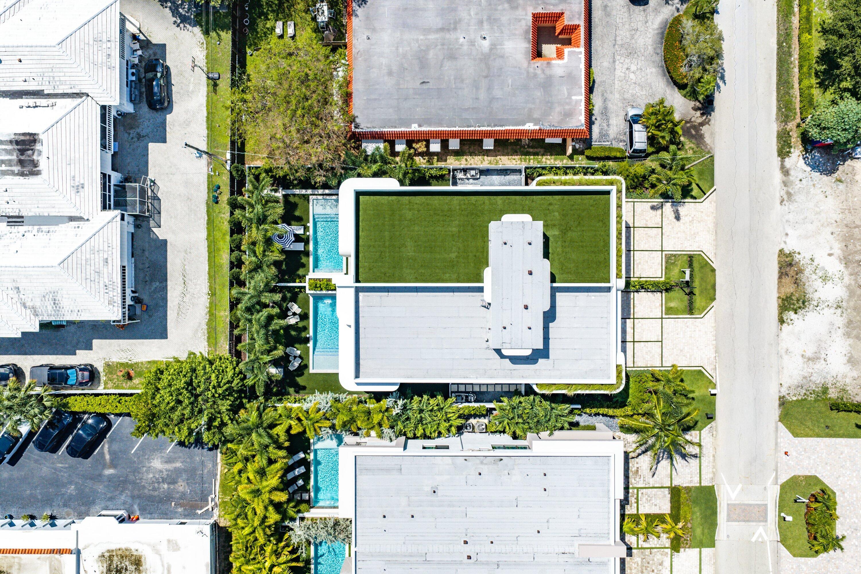 Aerial view of a modern home with a flat roof, turquoise pool, and palm-filled landscaping.