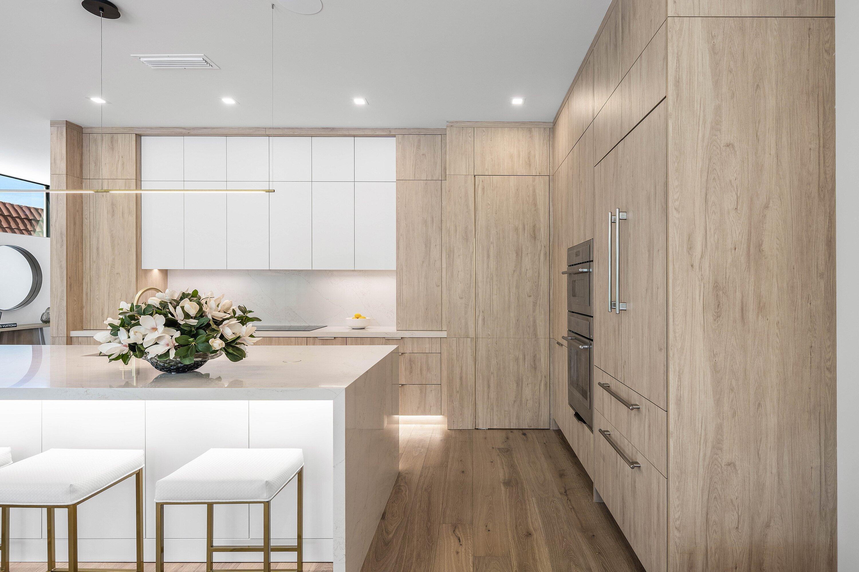 Modern kitchen with light wood cabinetry, a white stone island, built-in ovens, and upholstered bar stools.
