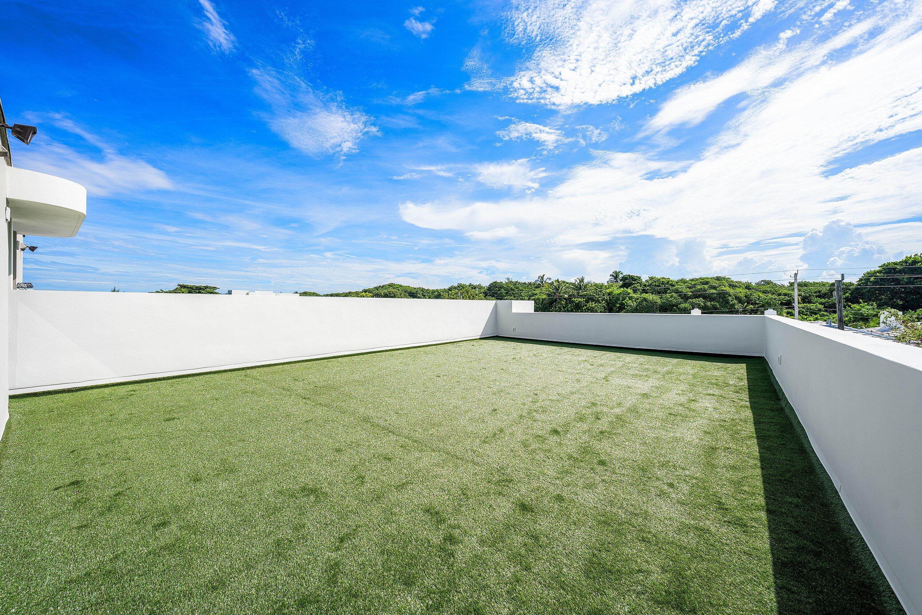 Rooftop terrace with green turf and low white walls beneath a bright, cloud-filled sky.