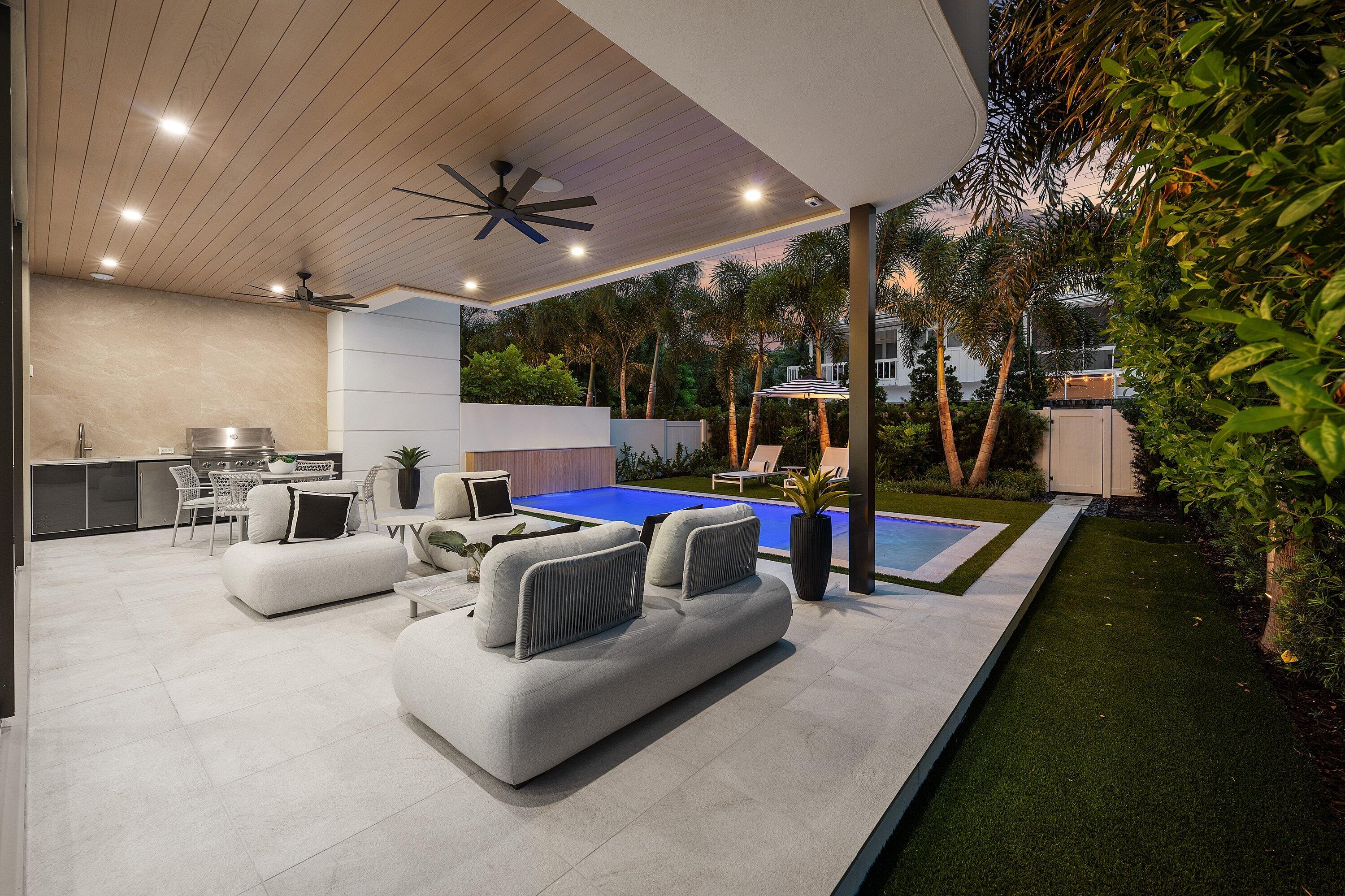 Covered patio with a built-in grill, lounge seating, swimming pool, and tropical landscaping at dusk.