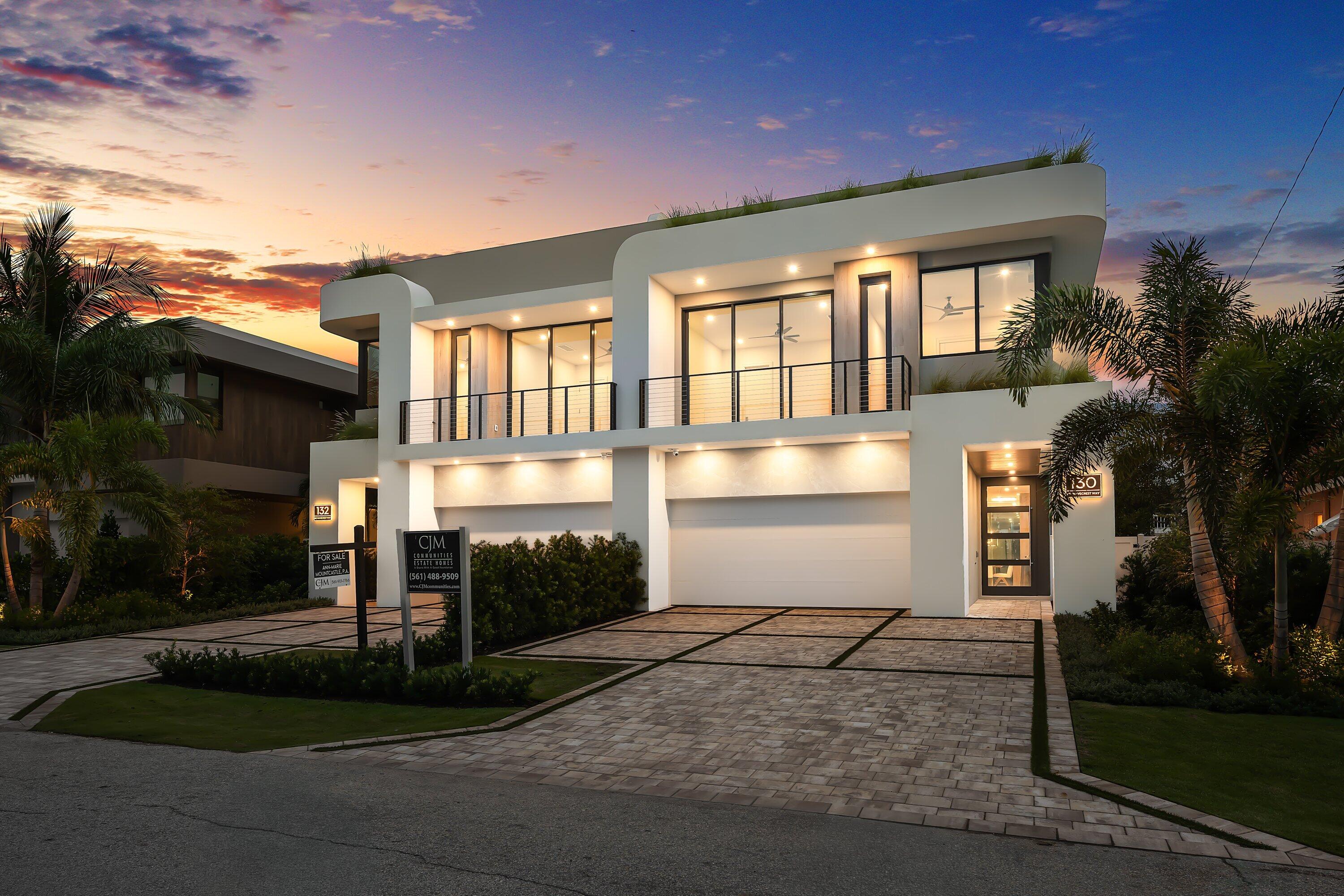 Modern two-story white home with a balcony and paved driveway, illuminated at dusk among palm trees.