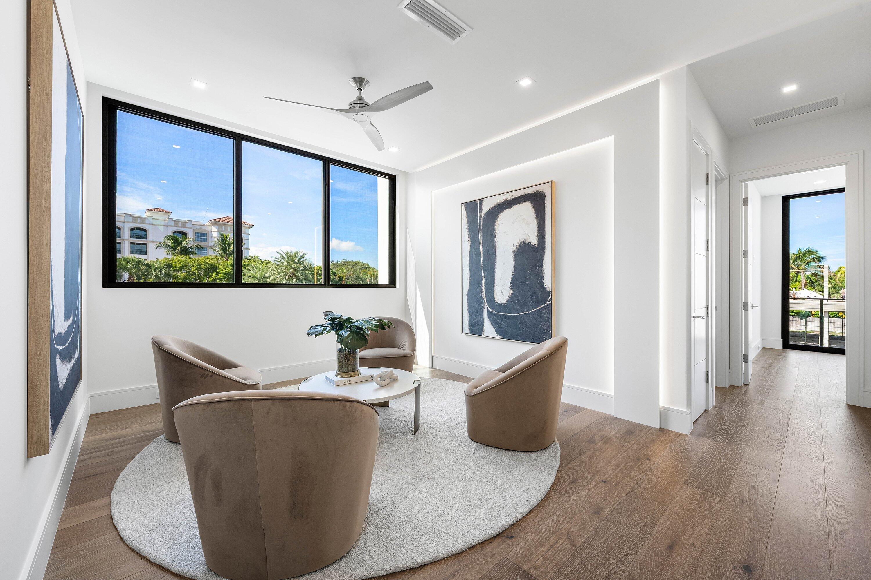 Furnished sitting room with taupe chairs, abstract art, and large windows overlooking palm trees.