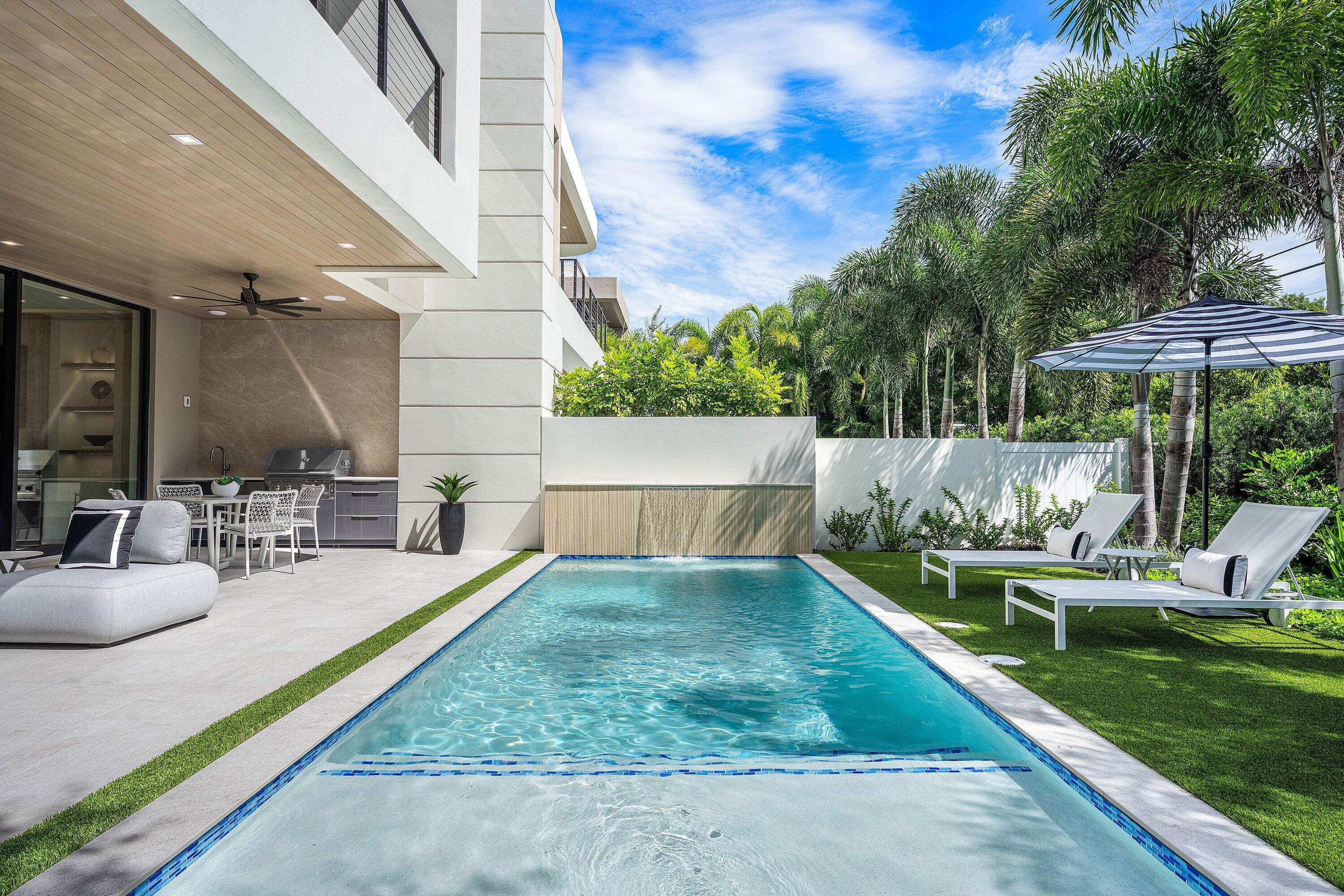 Modern home’s rectangular pool beside a furnished patio, built-in grill, and tropical landscaping.