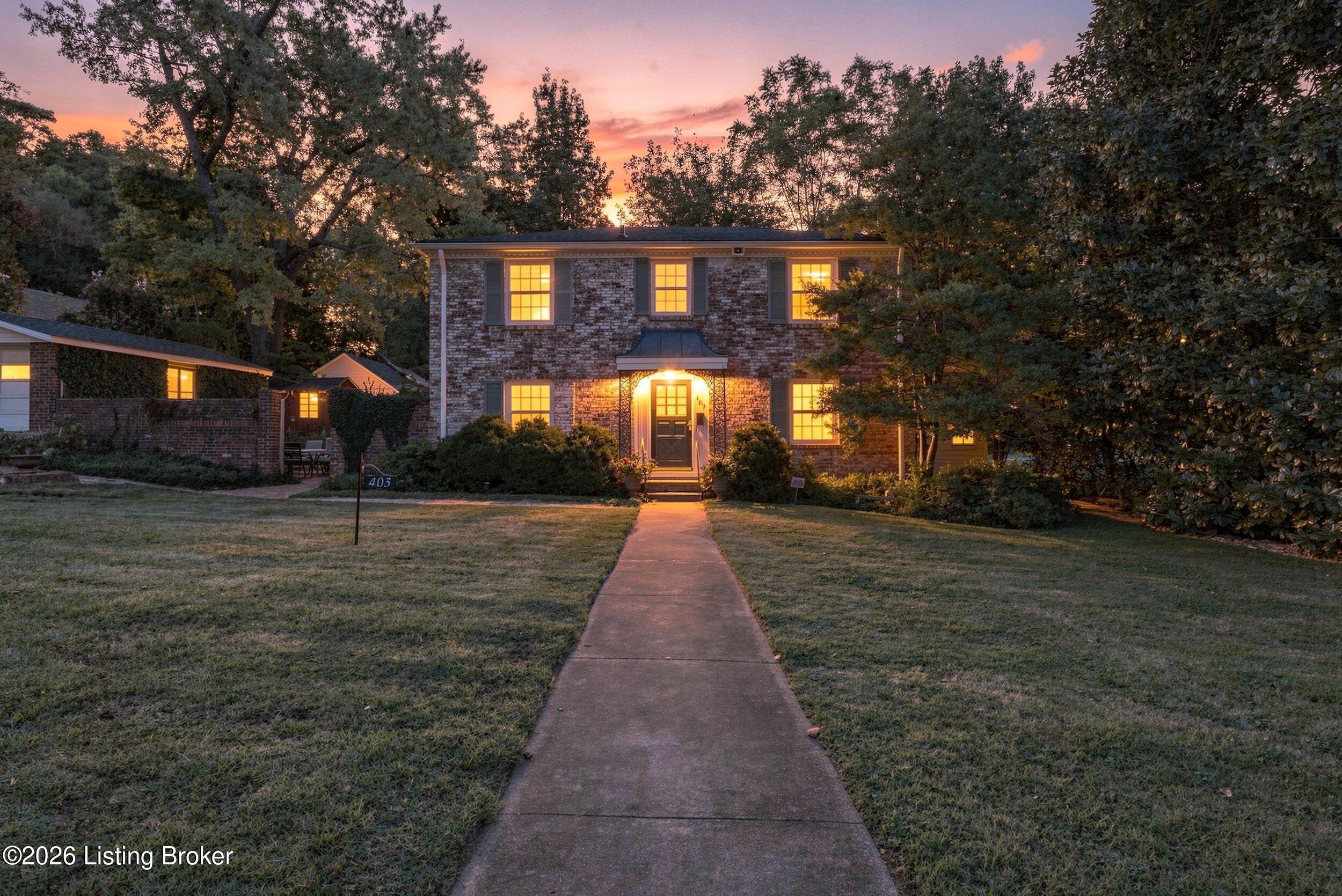Two-story brick home at dusk with glowing windows and a walkway through its tree-lined front yard.