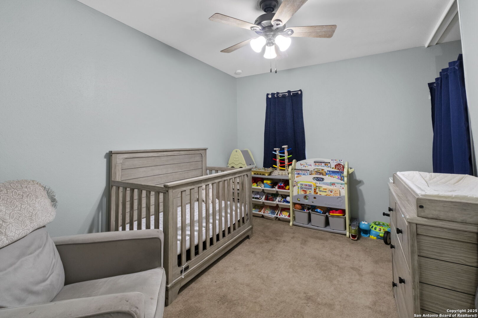 Nursery with a gray wooden crib, matching dresser, toy shelves, and blue curtains.