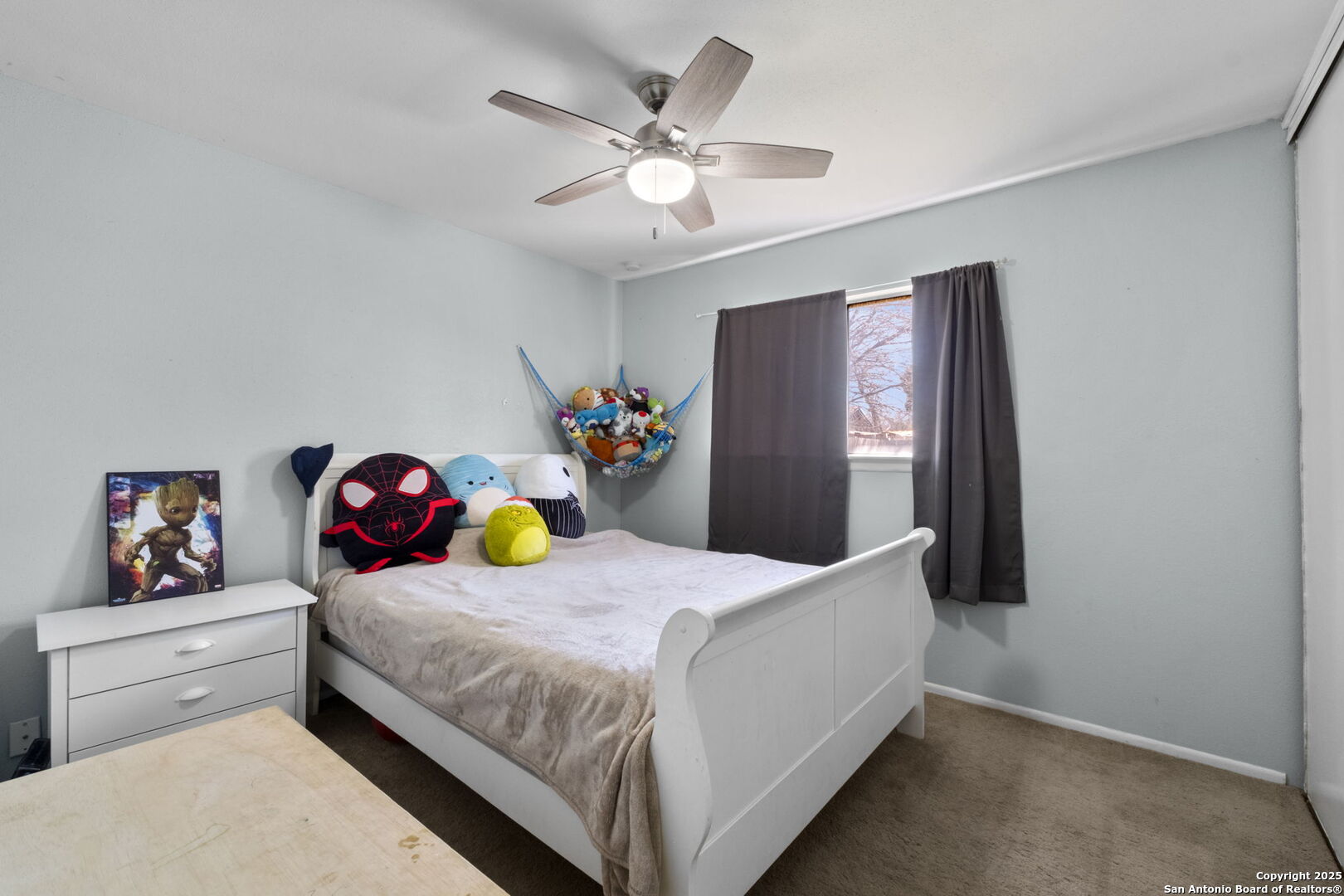 Bedroom with a white bed, colorful plush toys, a toy hammock, and a ceiling fan.