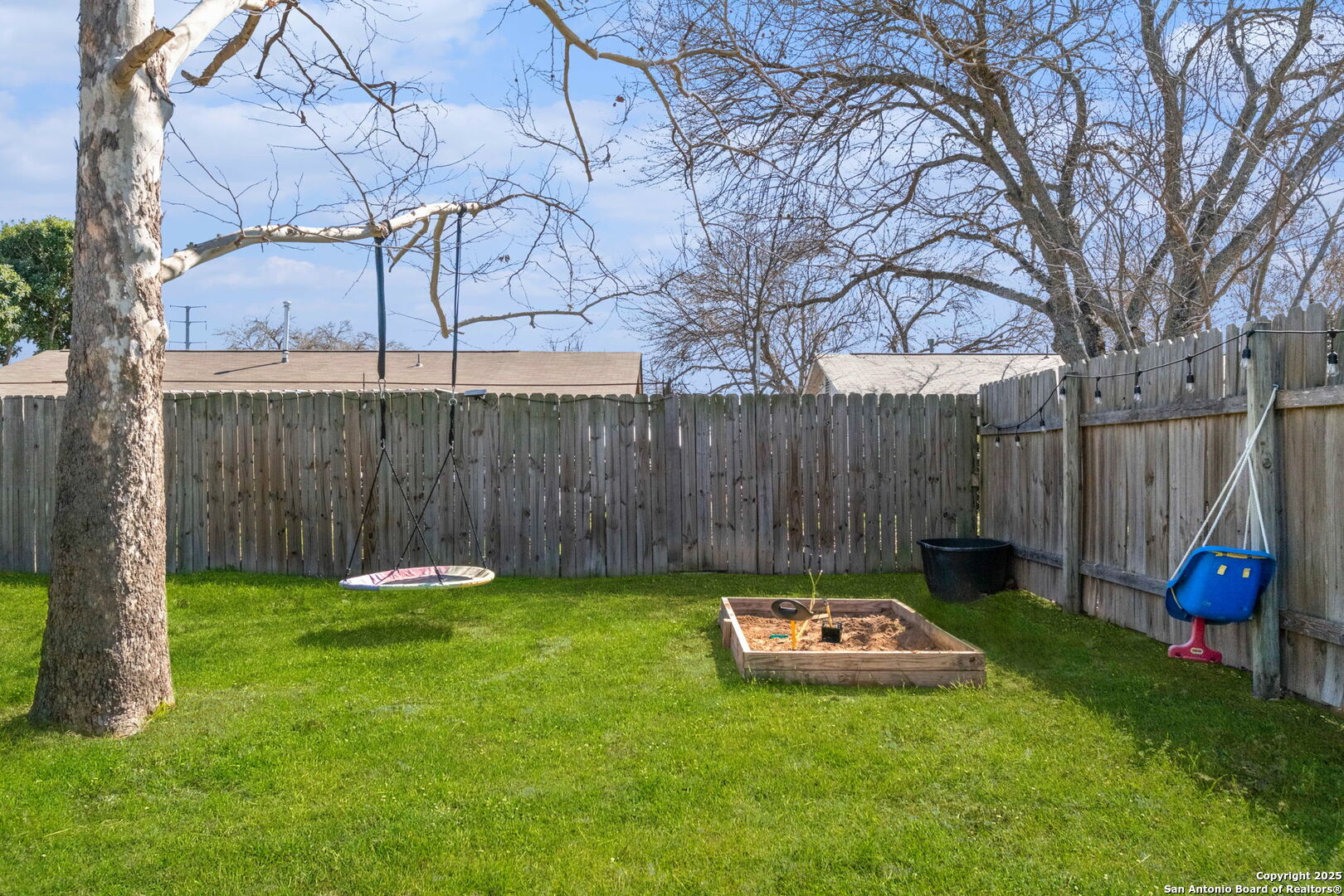 Fenced backyard with a green lawn, hanging swing, raised sandpit, and mature leafless trees.