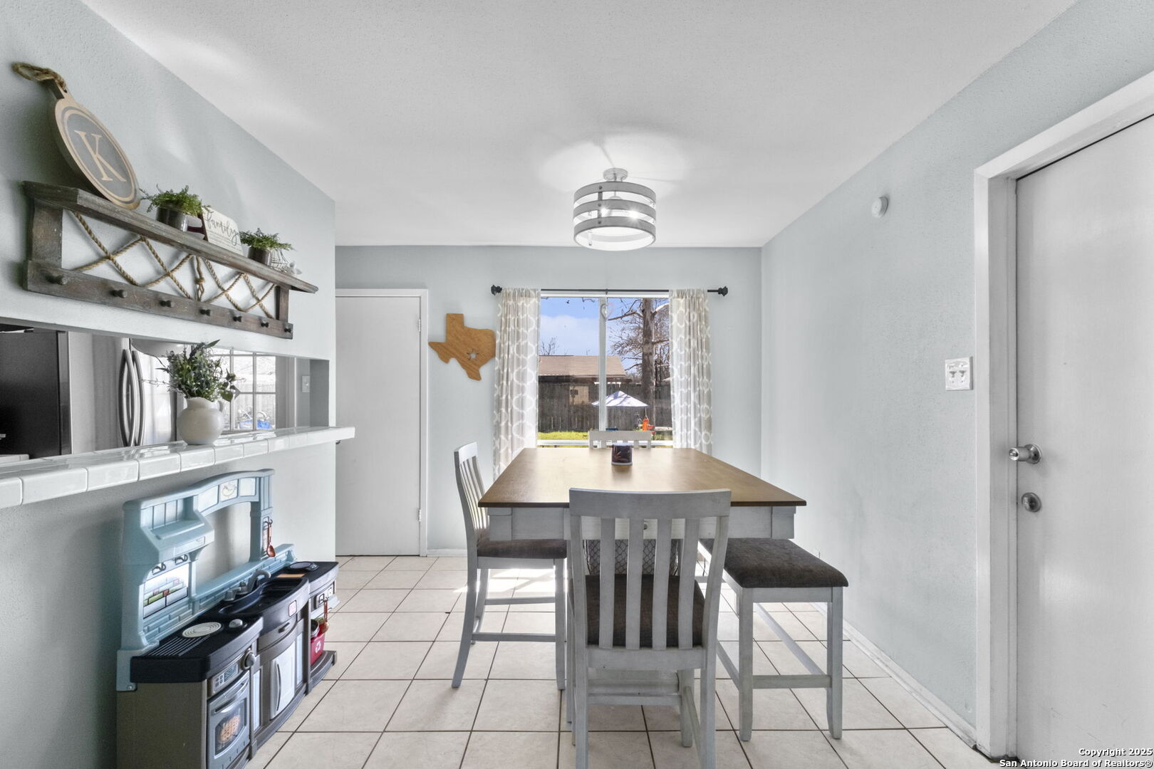 Dining room with a wooden table and gray seating beside a bright backyard-facing window.