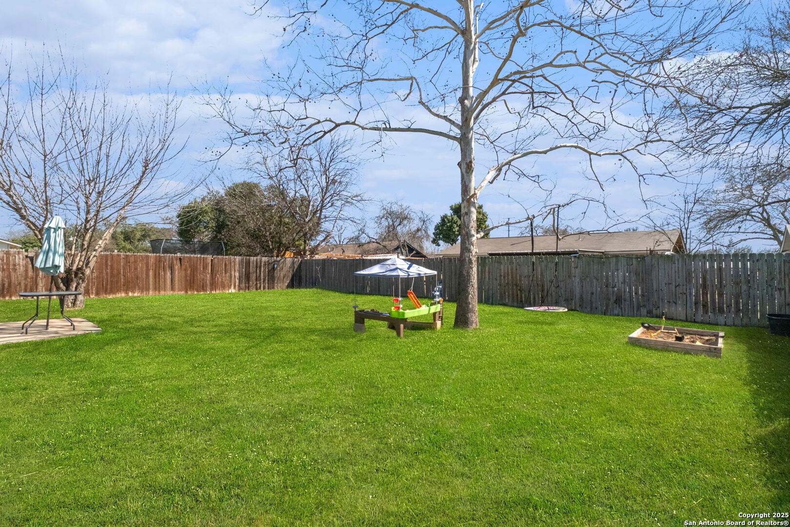 Fenced backyard with a broad green lawn, mature bare trees, and outdoor play equipment.