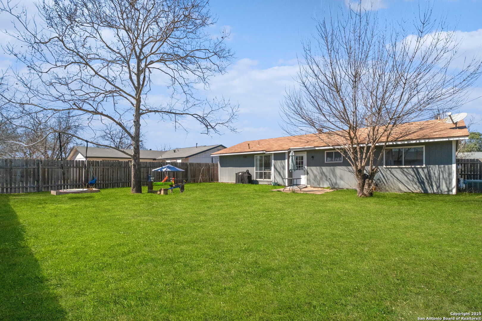 Fenced backyard with a broad green lawn, two leafless trees, a small patio, and a children’s play area.