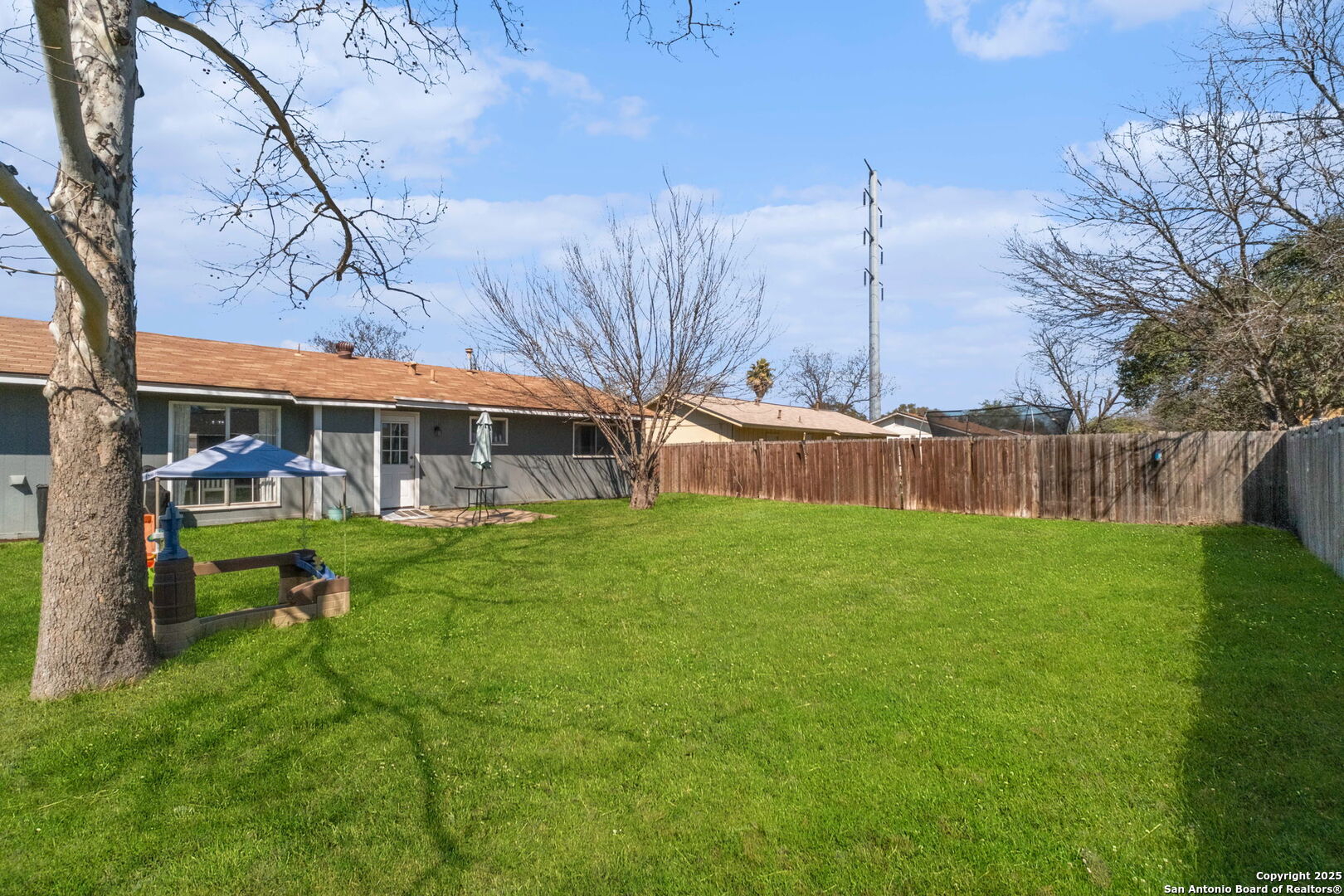Fenced backyard with a broad green lawn, mature trees, and a patio behind a single-story gray home.