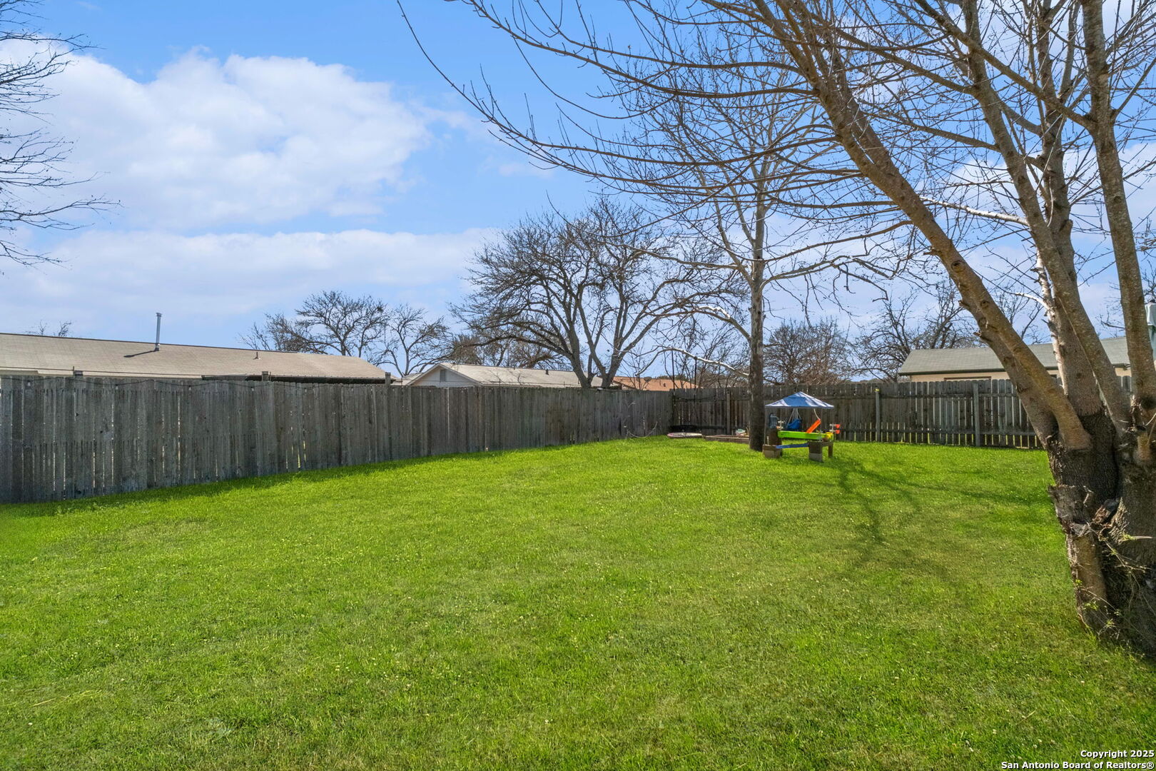 Fenced backyard with a broad green lawn, mature trees, and a small children’s play area.