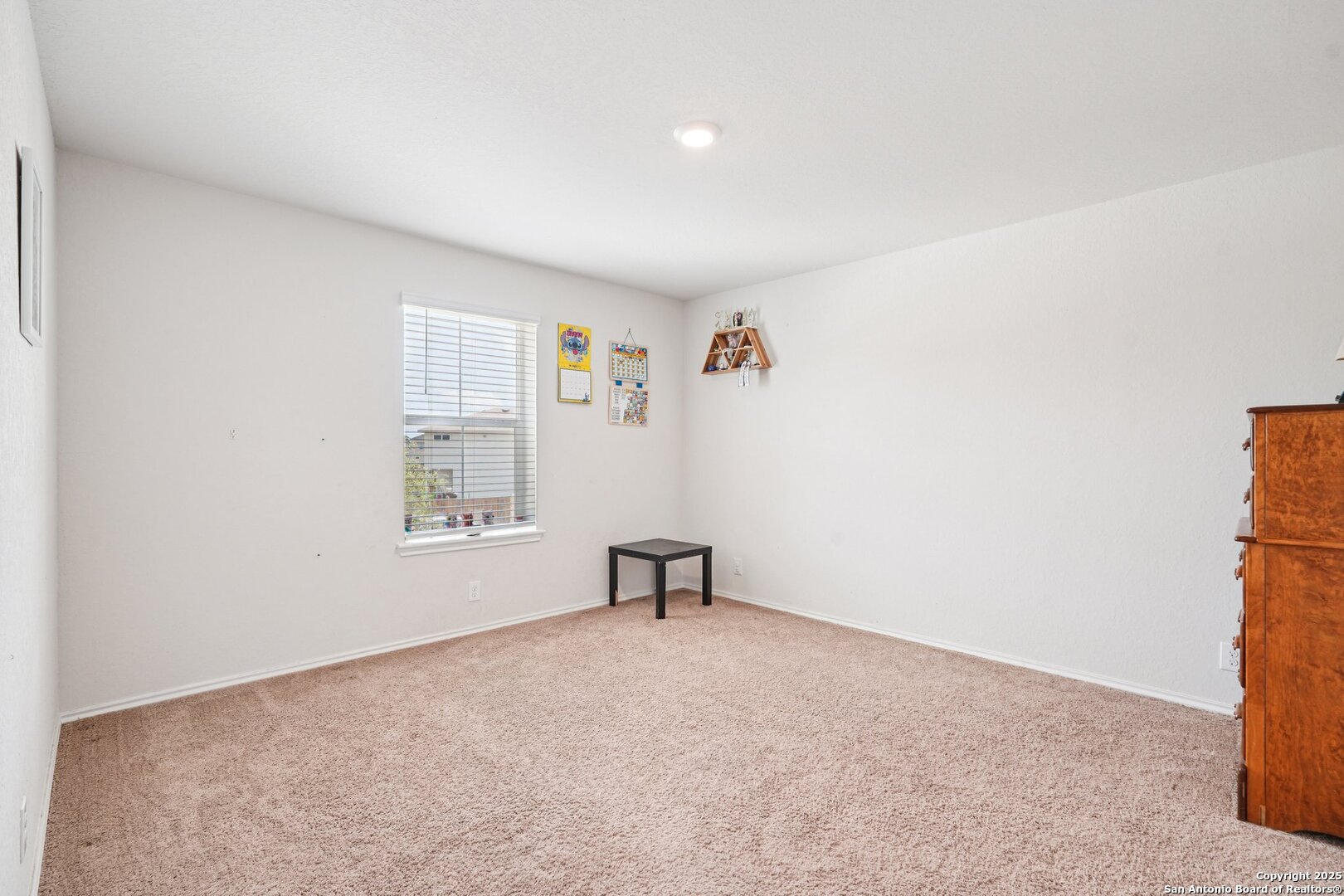 Carpeted bedroom with a window, small table, wall calendars, and wooden dresser.