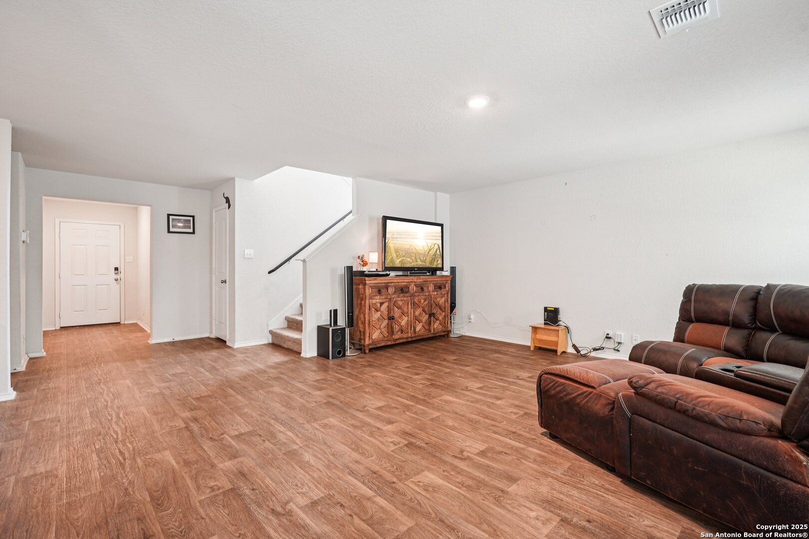 Living room with a brown leather sectional, wood-look floors, and a media cabinet beside the staircase.