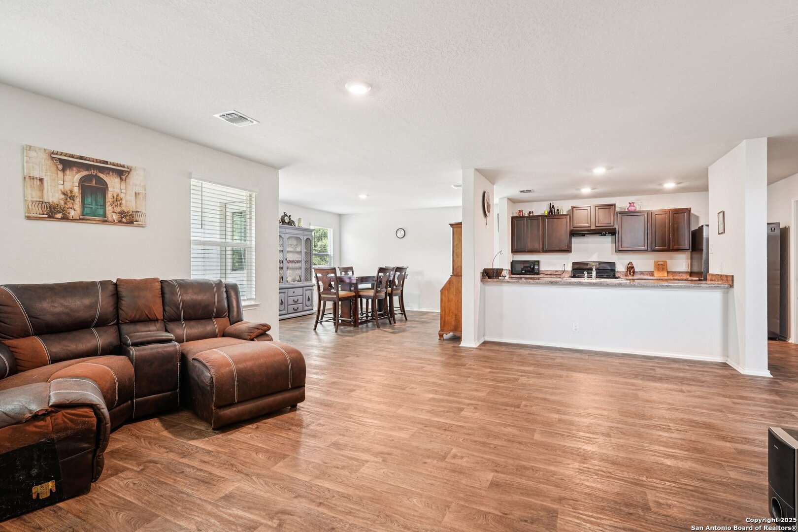 Open-plan living room, dining area, and kitchen with a brown leather sectional and wood-look flooring.