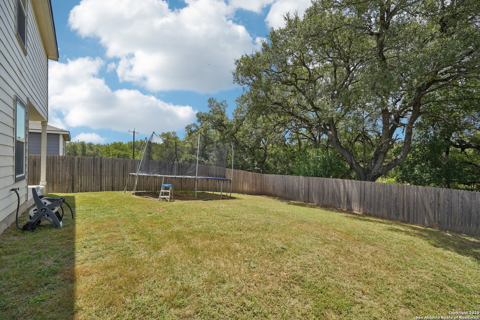 Fenced backyard with a trampoline, grassy lawn, and mature trees.