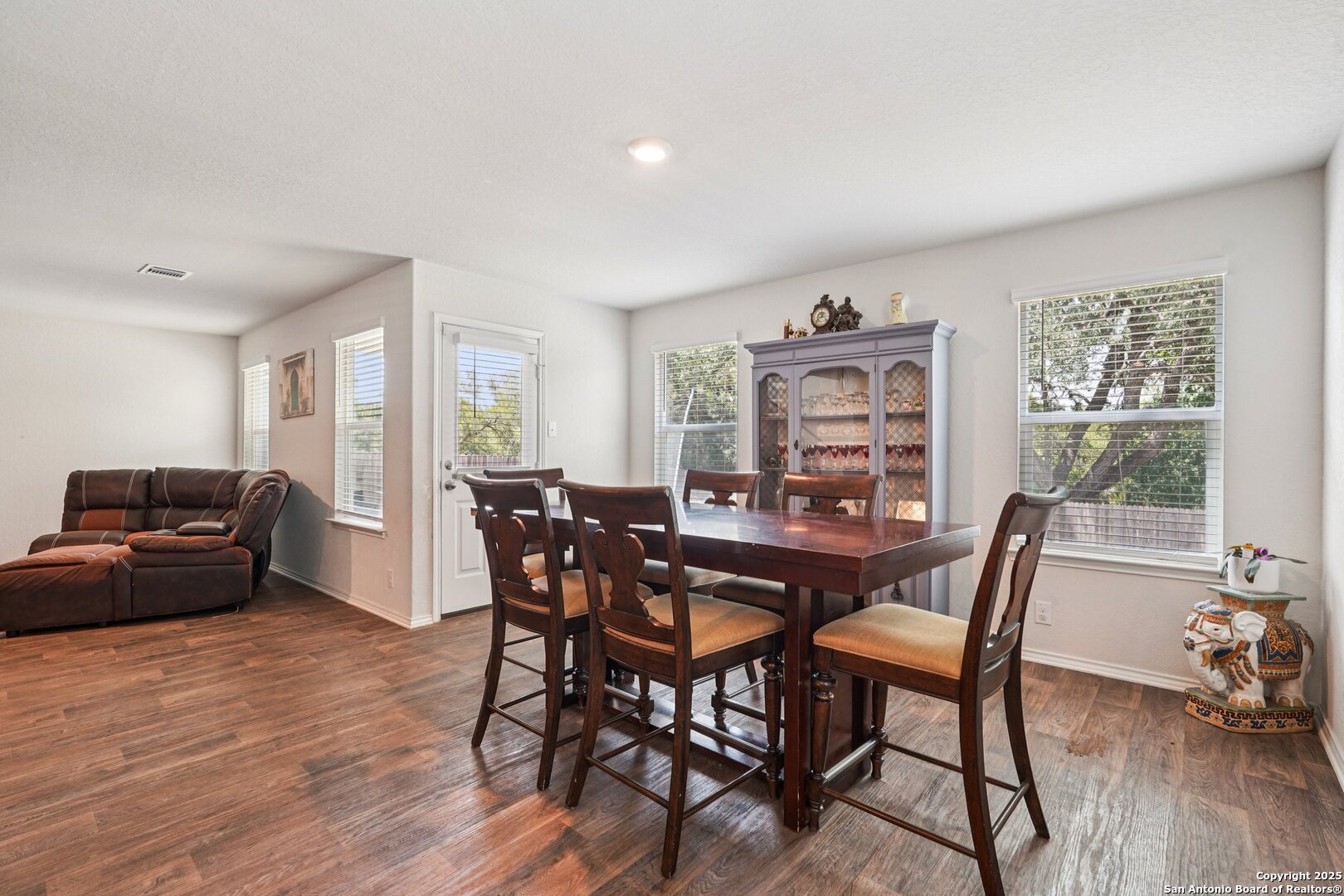 Dining area with a dark wood table and chairs beside a glass-front china cabinet and leather sectional.
