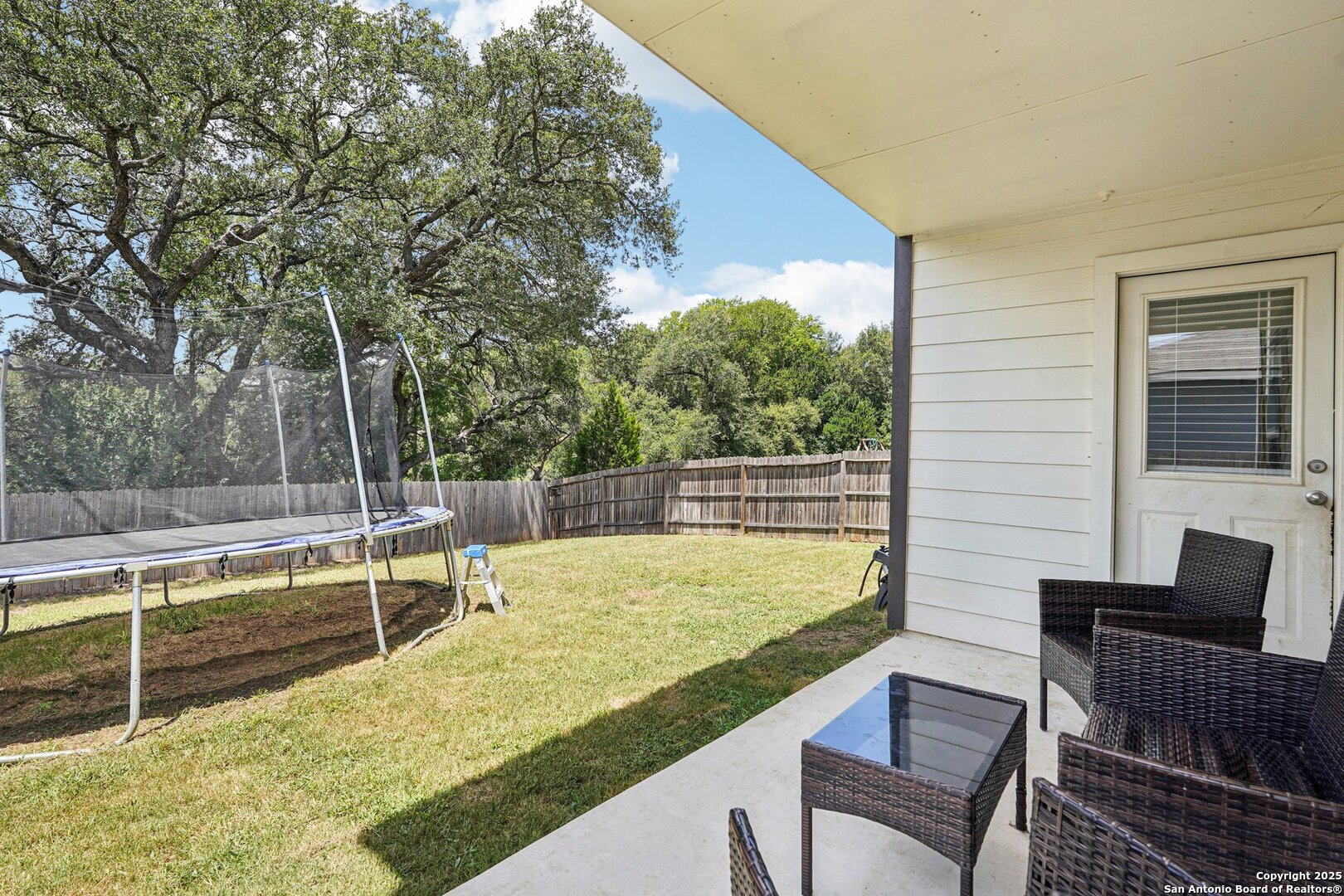 Covered patio overlooking a fenced grassy backyard with wicker furniture and a large trampoline.
