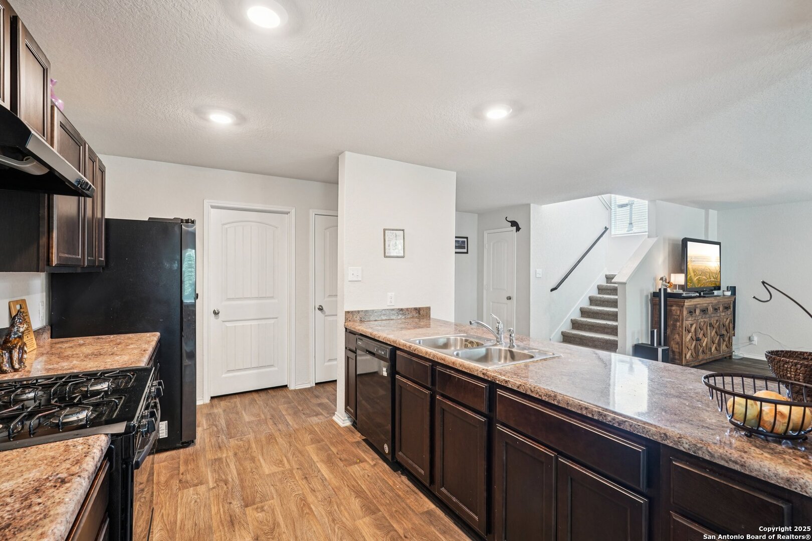 Kitchen with dark cabinets, stone counters, a double sink, and an open view of the staircase.