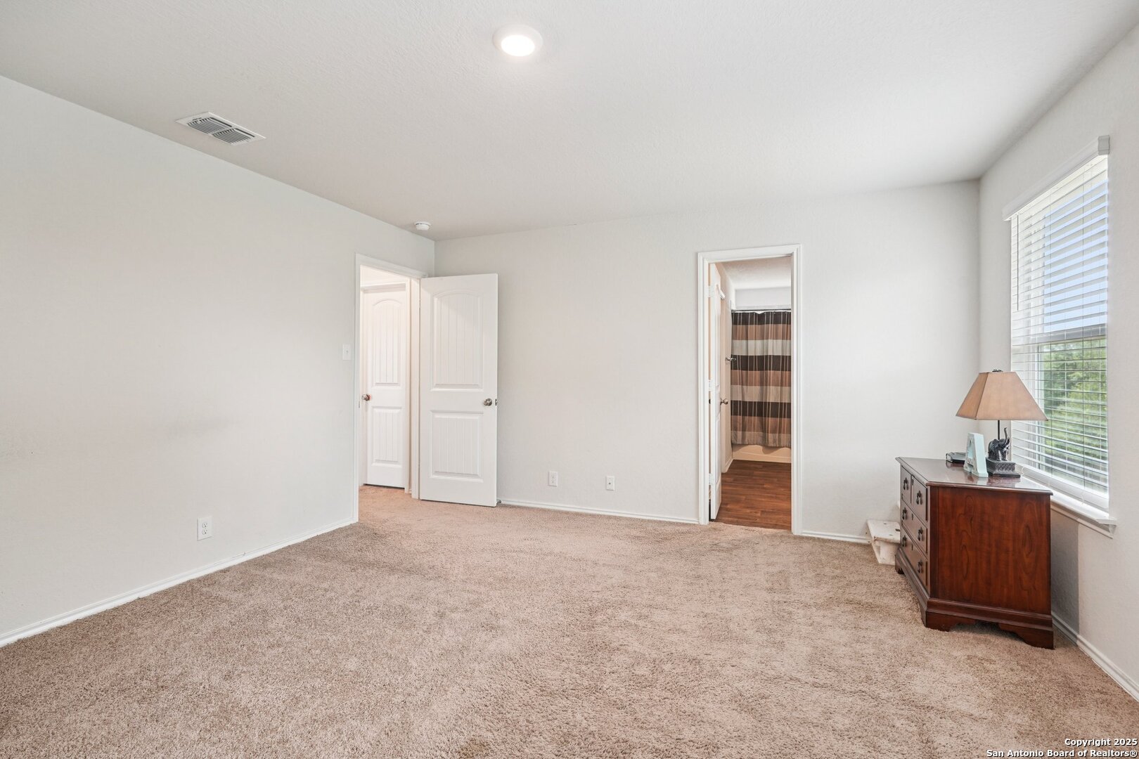 Carpeted bedroom with a wooden dresser by the window and an adjoining bathroom.