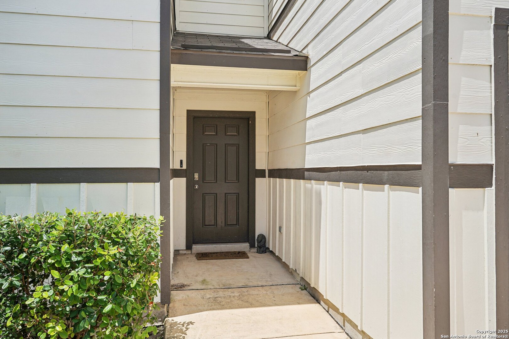 Recessed dark front door beneath a covered entry, framed by white siding and a leafy shrub.
